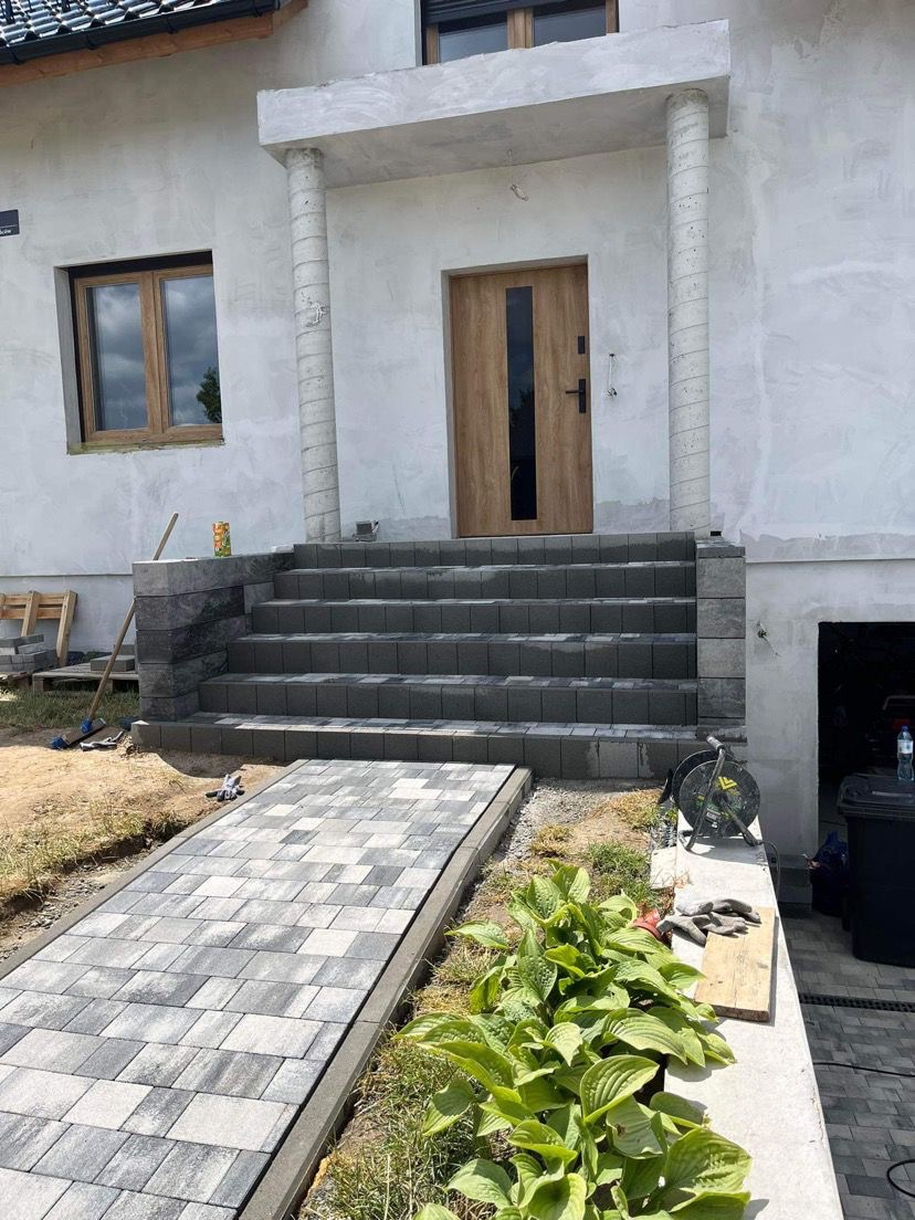Ułożony chodnik z szaro-białej kostki prowadzi do schodów wejściowych domu z drewnianymi drzwiami; świeżo posadzone rośliny ozdabiają otoczenie chodnika.