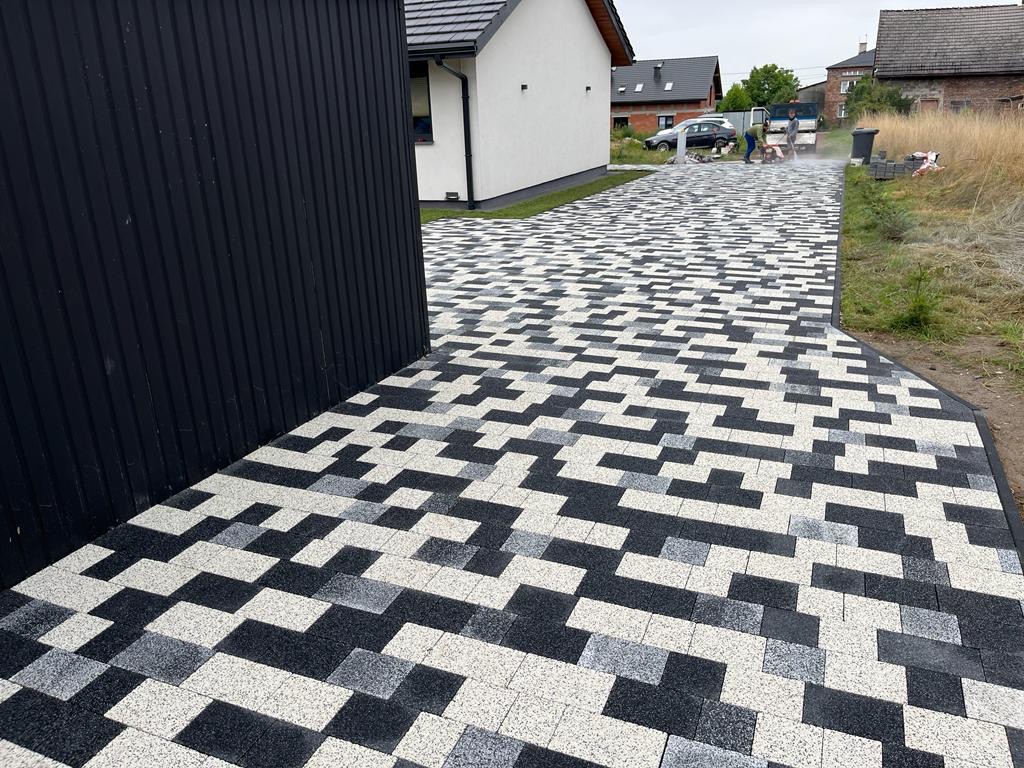 Nowo ułożony podjazd z kostki brukowej w odcieniach szarości i bieli, tworzący geometryczny wzór, widok z perspektywy niskiego kąta, z fragmentem czarnej elewacji budynku po lewej stronie.