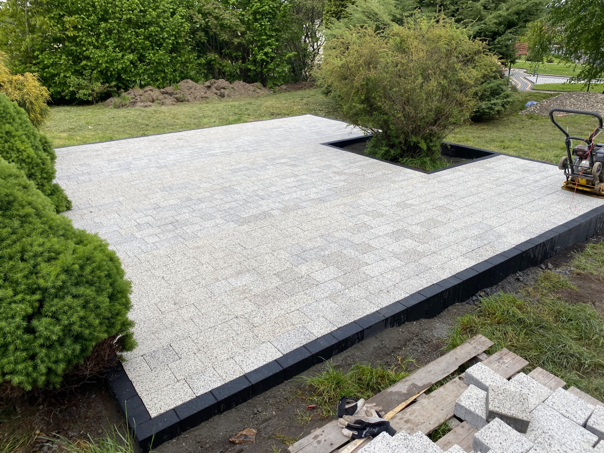 Świeżo ułożony, jasnoszary plac z kostki brukowej z czarnym obramowaniem, częściowo otaczający rabatę z krzewami. Widoczna zagęszczarka w tle i paleta z kostką w rogu.