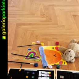 Jasny parkiet ułożony we wzór jodełki francuskiej, z rozłożonymi farbami, pędzlami i rysunkiem dziecka przedstawiającym misia, obok leży pluszowy miś.