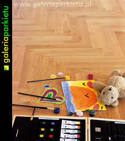 Jasny parkiet ułożony we wzór jodełki francuskiej, z rozłożonymi farbami, pędzlami i rysunkiem dziecka przedstawiającym misia, obok leży pluszowy miś.