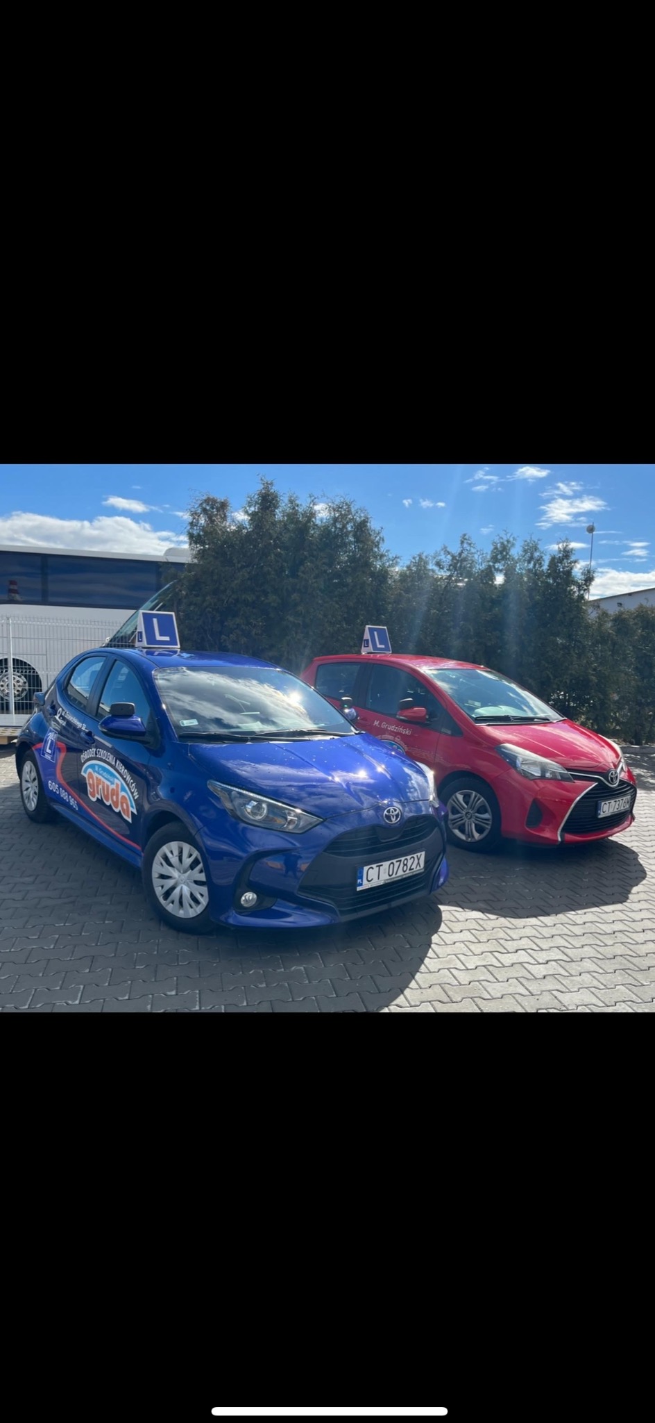 Dwa samochody Toyota Yaris, niebieski i czerwony, z oznaczeniami 'L' na dachu i naklejkami z nazwą firmy na boku, stojące na parkingu z kostki brukowej w słoneczny dzień.