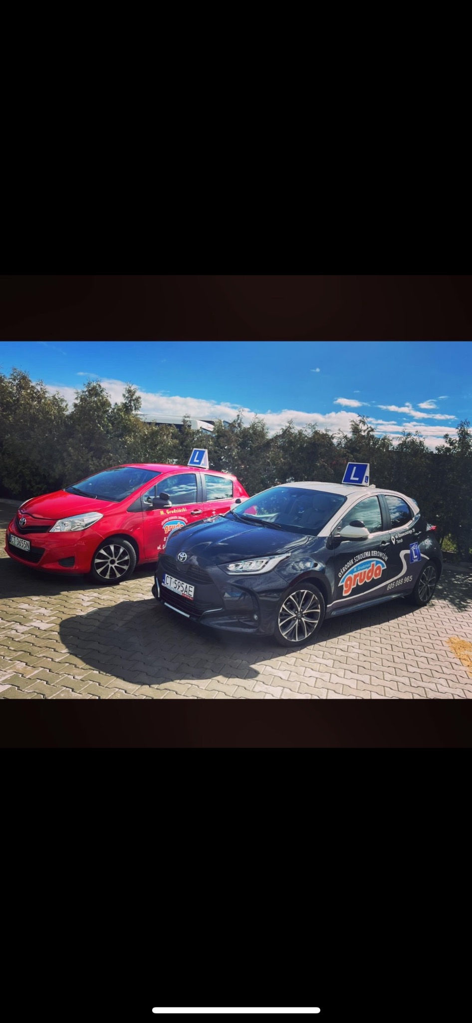 Dwa samochody Toyota Yaris, czerwony i czarny, z oznaczeniami 'L' na dachu, zaparkowane na brukowanym placu przed zielenią, należące do szkoły jazdy 'Gruda' z Torunia, widoczne numery rejestracyjne.