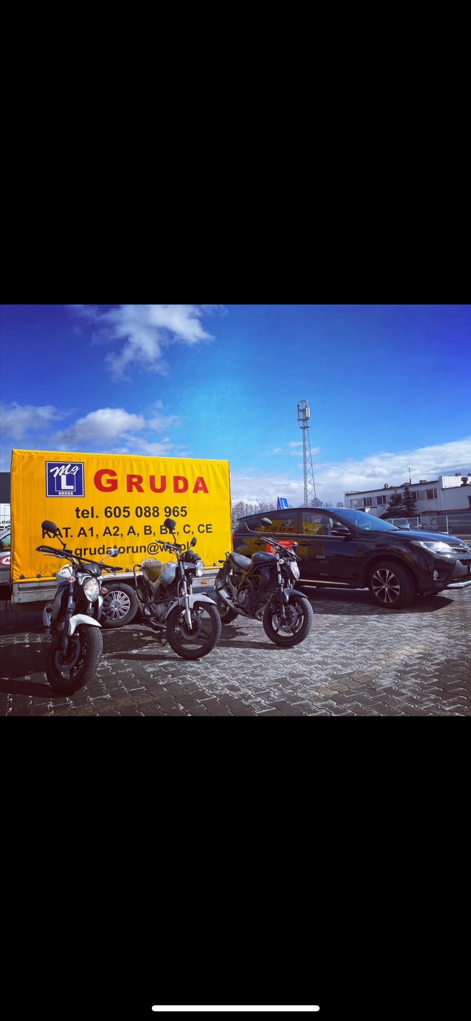 Trzy motocykle przed żółtą przyczepą z logo i danymi kontaktowymi, zaparkowane na kostce brukowej, w tle czarny SUV, błękitne niebo z chmurami i maszt telekomunikacyjny.