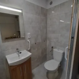 Wykończona łazienka z szarymi płytkami, umywalką na drewnianej szafce, toaletą i uchwytem dla niepełnosprawnych, widoczne elementy instalacji elektrycznej i wentylacja.