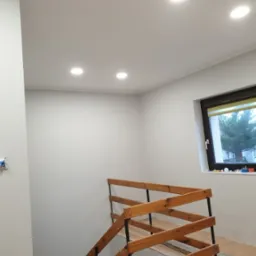 Korytarz w domu jednorodzinnym po remoncie: świeżo pomalowane ściany w odcieniach szarości, drewniane schody z czarnymi elementami metalowymi, zabezpieczone podłogi i okno z widokiem na zieleń.