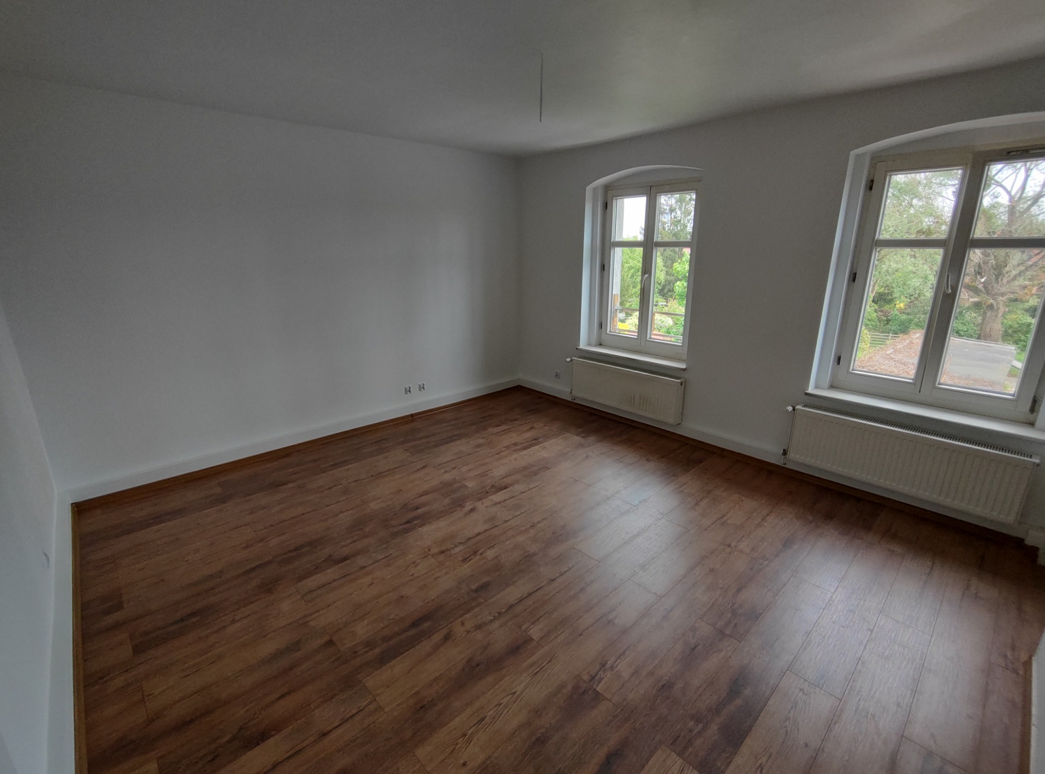 Pusty pokój z panelami podłogowymi w odcieniach brązu, świeżo pomalowanymi białymi ścianami i dwoma oknami z widokiem na zieleń.