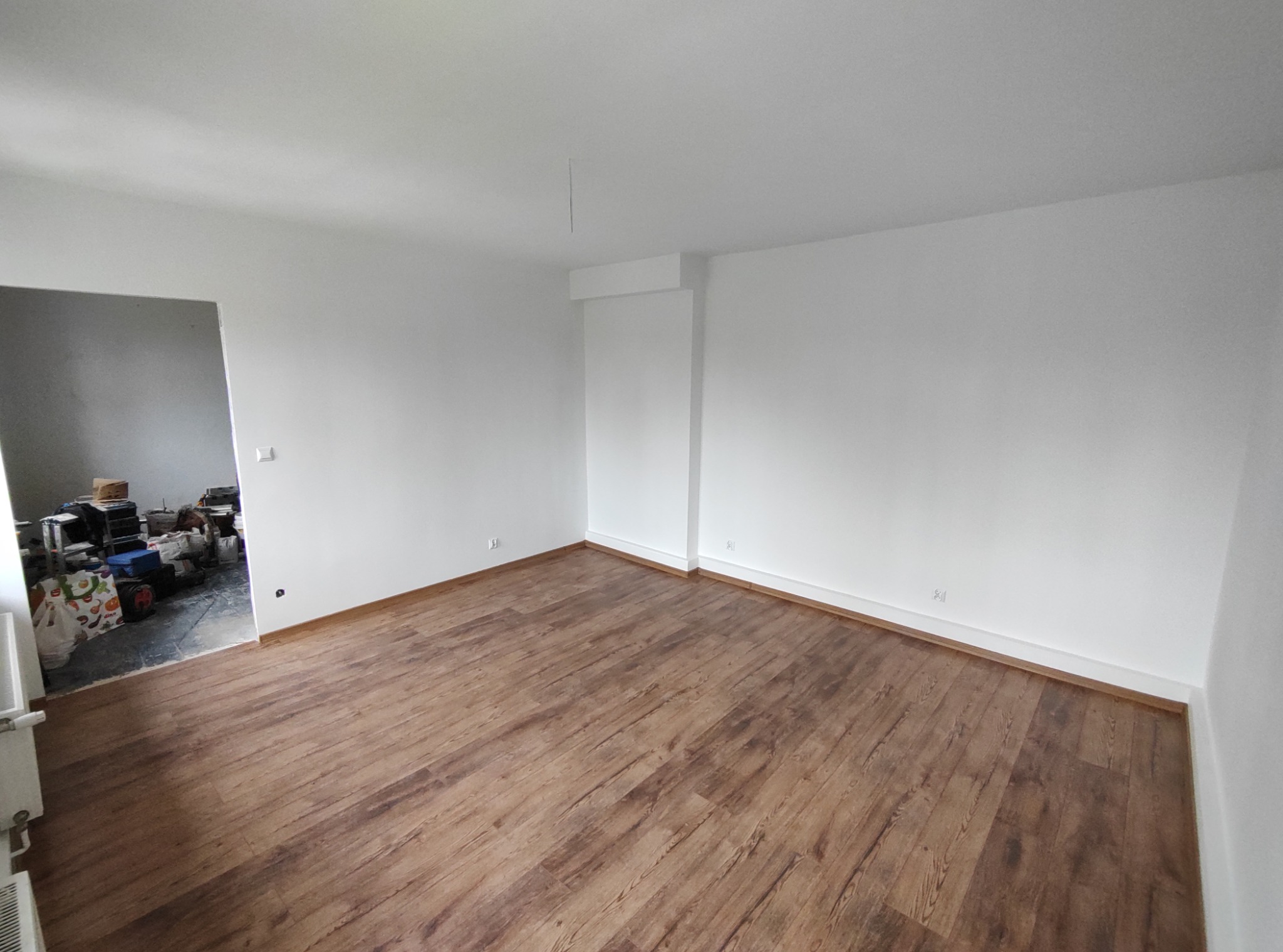 Puste, odnowione pomieszczenie z brązowymi panelami podłogowymi imitującymi drewno, świeżo pomalowanymi białymi ścianami i widocznym bałaganem w sąsiednim pokoju.