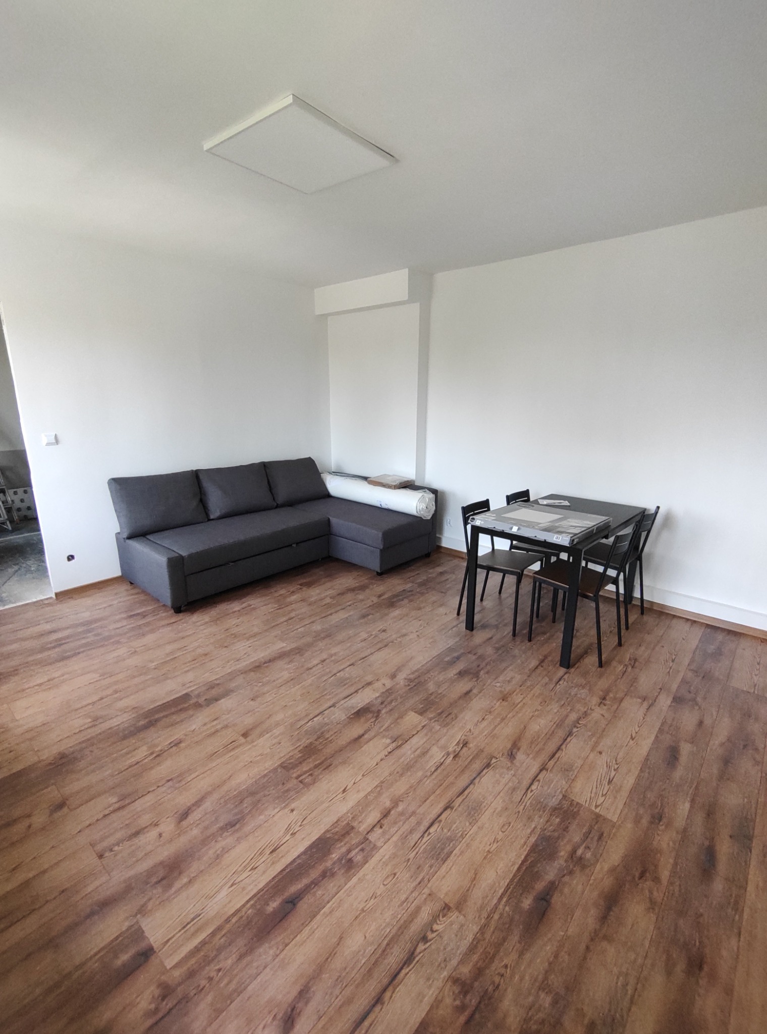 Salon z drewnopodobną podłogą, szarą sofą narożną, czarnym stołem i krzesłami, oraz panelem LED na suficie.