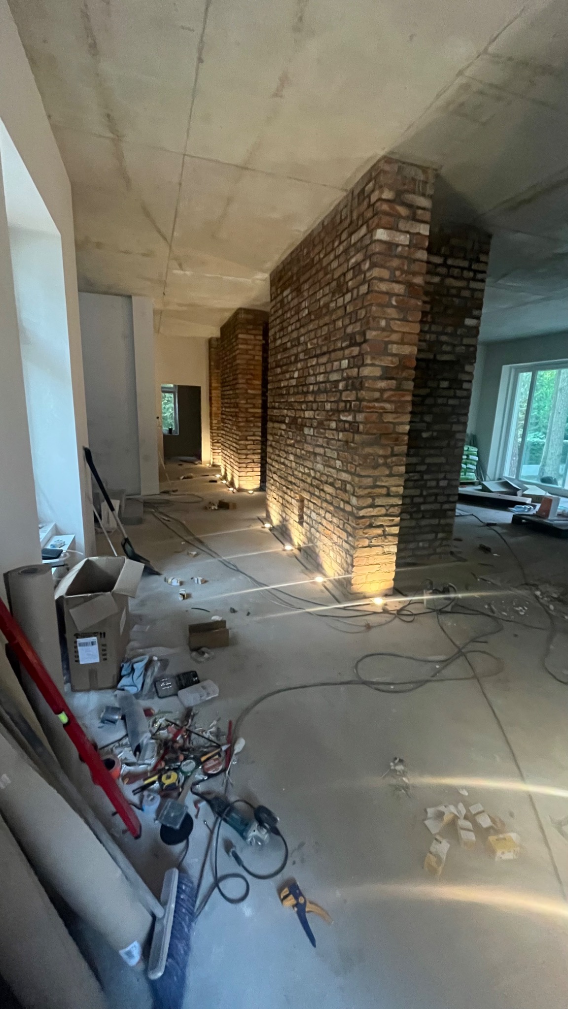 Wnętrze w trakcie remontu z widocznymi dwoma kolumnami z cegły, surowym betonowym sufitem i przewodami elektrycznymi wystającymi z podłogi, oświetlonymi punktowymi lampami podłogowymi.