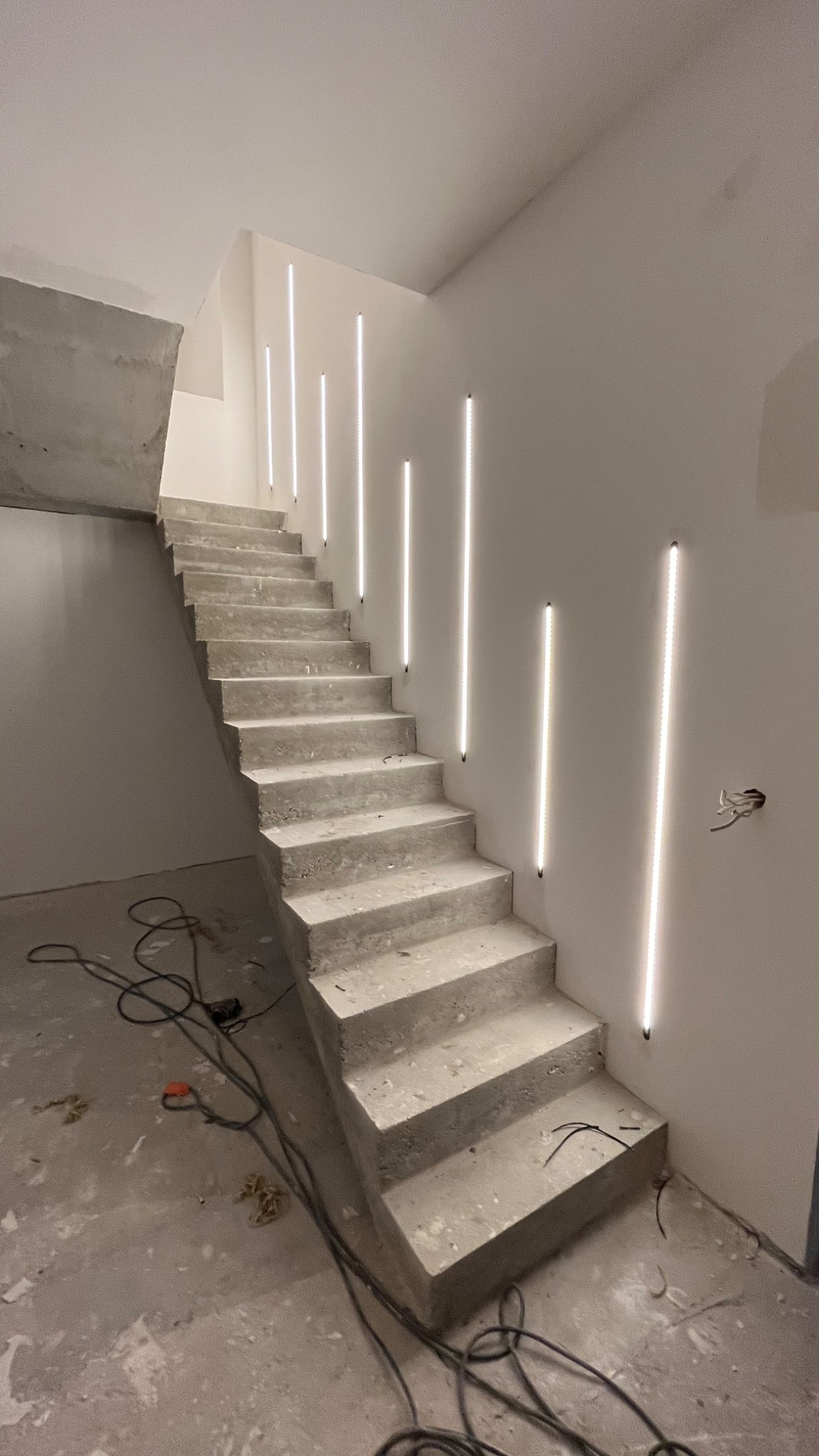 Surowe betonowe schody z nowoczesnym oświetleniem LED w postaci pionowych pasków na ścianie, widoczne kable i niedokończone wykończenie wnętrza.