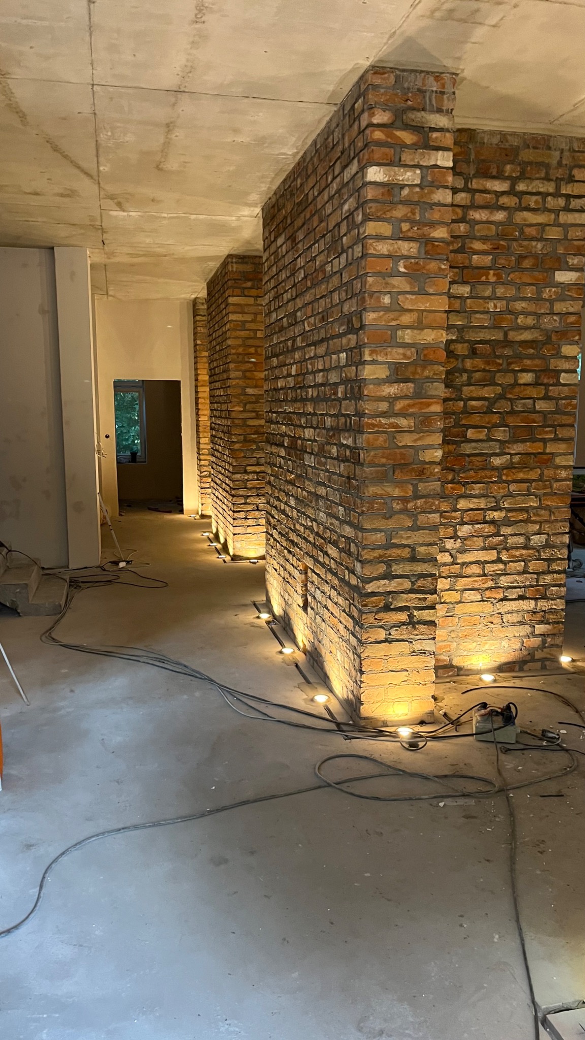 Surowy stan wykończenia wnętrza z ceglanymi filarami oświetlonymi punktowymi lampami podłogowymi, widoczne kable i narzędzia na posadzce.
