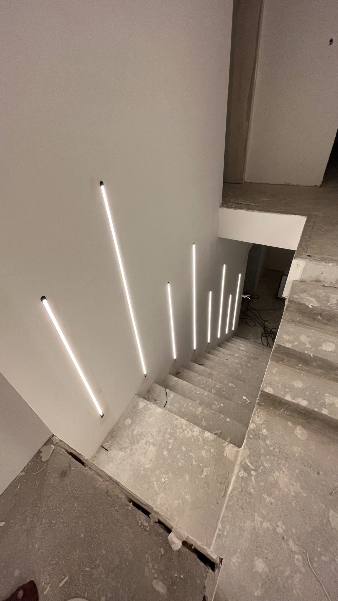 Widok z góry na betonowe schody w trakcie wykończenia z zamontowanym pionowym oświetleniem LED na ścianie. Światło z lamp oświetla schody. Widoczne kable i niedokończone wykończenie.