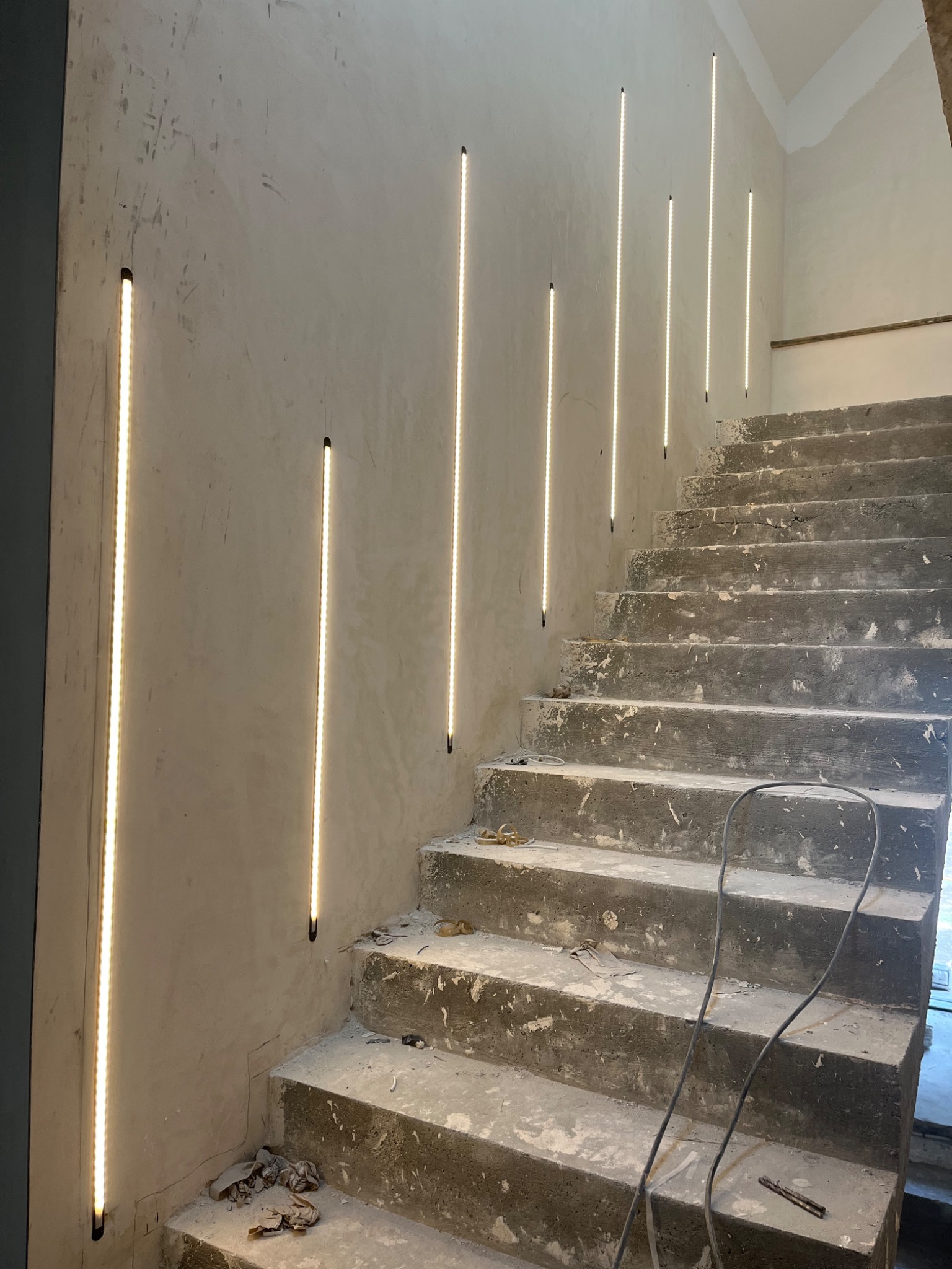 Surowe betonowe schody z pionowymi paskami ledowego oświetlenia w ścianie, widoczne przewody i narzędzia na stopniach.