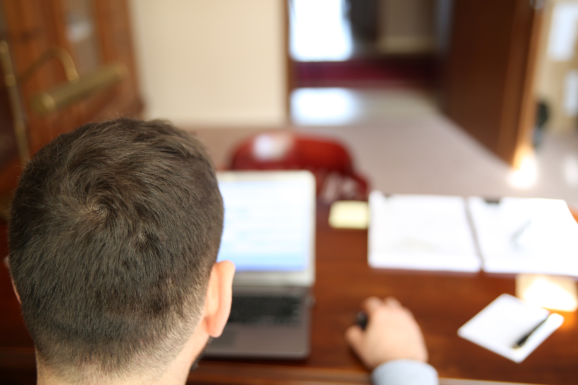 Mężczyzna siedzi przy biurku z laptopem, notatnikiem i długopisem, prawdopodobnie pracuje zdalnie lub analizuje dokumenty.