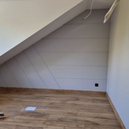 Sławek Grzesikiewicz - Pokój na poddaszu po malowaniu, z drewnianą podłogą, skośnymi ścianami z dekoracyjnymi listwami w kolorze szarym i białym sufitem, widoczny fragment osoby sprzątającej podłogę.