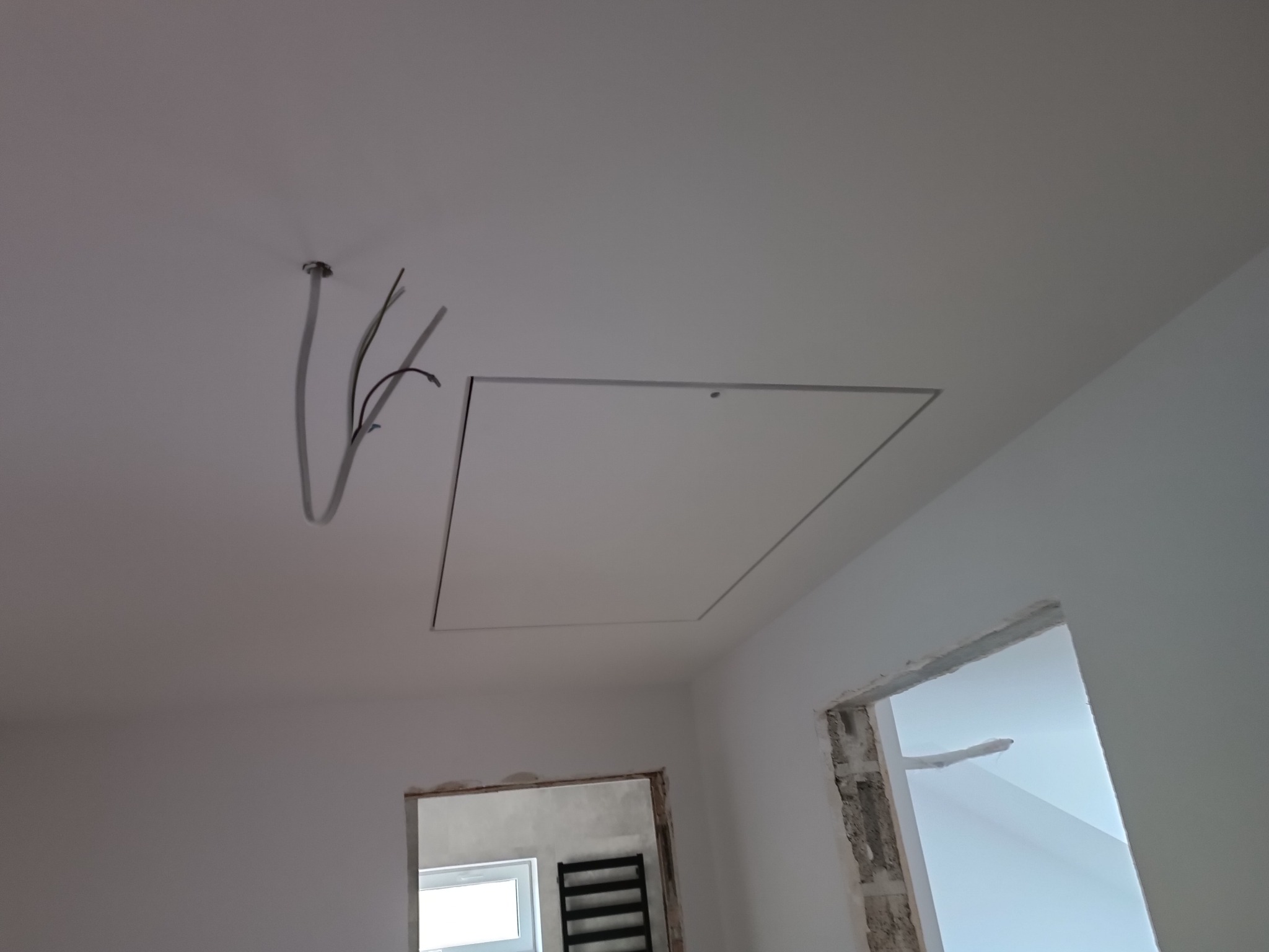 Białe wnętrze w trakcie remontu: widoczne kable wystające z sufitu, klapa rewizyjna w suficie, ściany po nałożeniu gładzi, oraz otwory drzwiowe w stanie surowym.