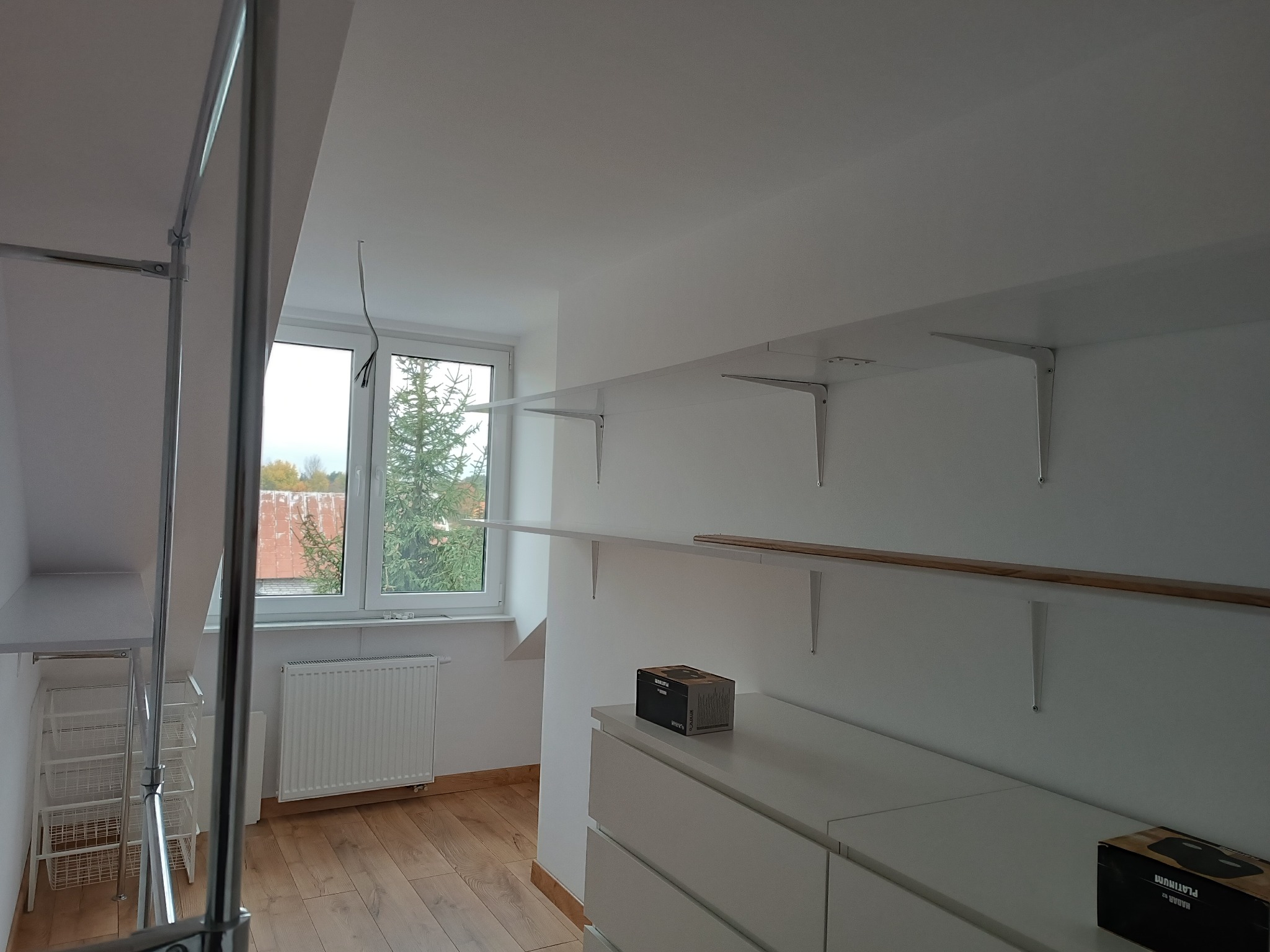 Biała garderoba w skosie poddasza z półkami na wspornikach, szufladami i koszami drucianymi, widok na okno z grzejnikiem.