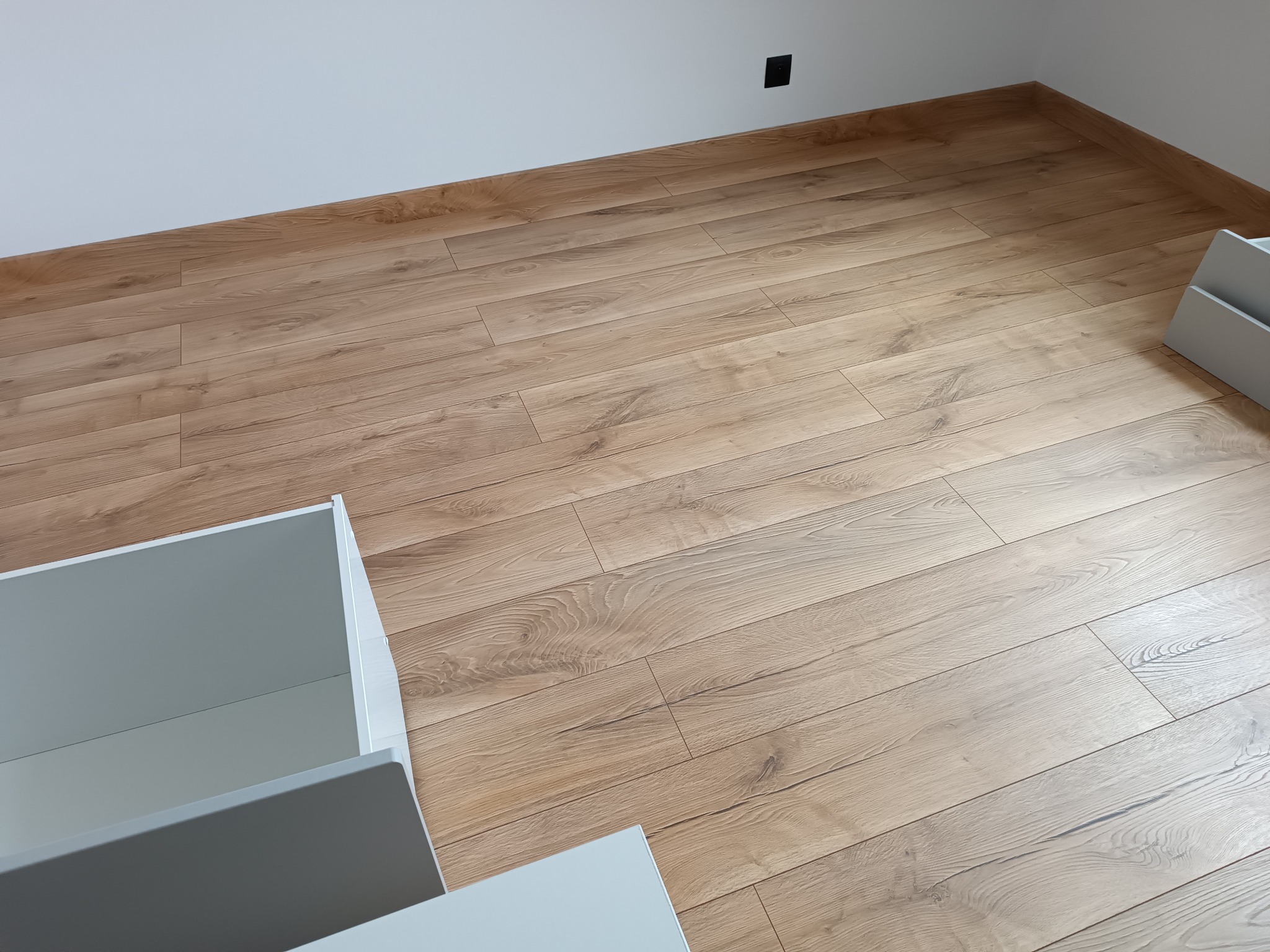 Ułożona podłoga panelowa w odcieniach jasnego drewna, widoczny fragment białej ściany z czarnym gniazdkiem elektrycznym oraz elementy białych mebli z szufladami.