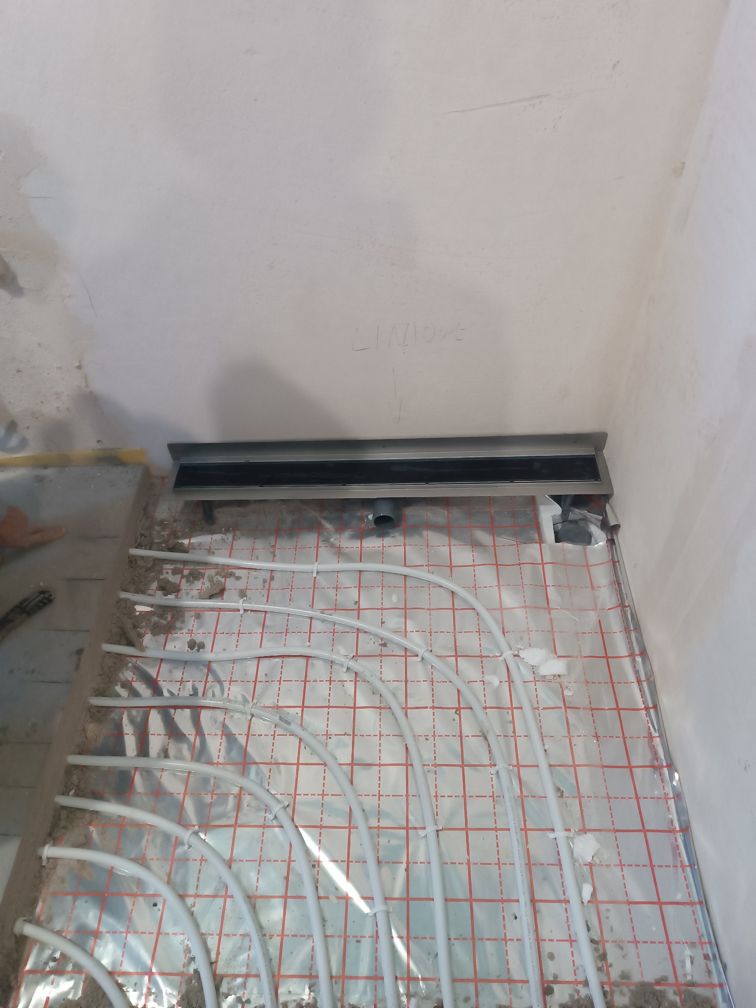 Ułożone rury ogrzewania podłogowego na folii z siatką, odpływ liniowy w rogu łazienki, ściany w stanie surowym.