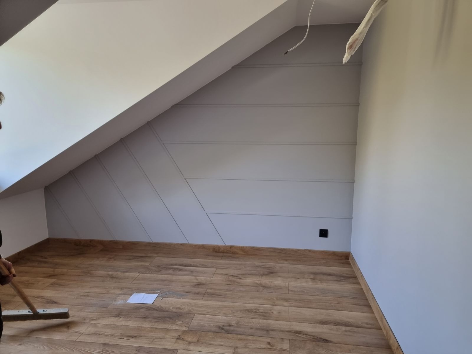 Pokój na poddaszu po malowaniu, z drewnianą podłogą, skośnymi ścianami z dekoracyjnymi listwami w kolorze szarym i białym sufitem, widoczny fragment osoby sprzątającej podłogę.