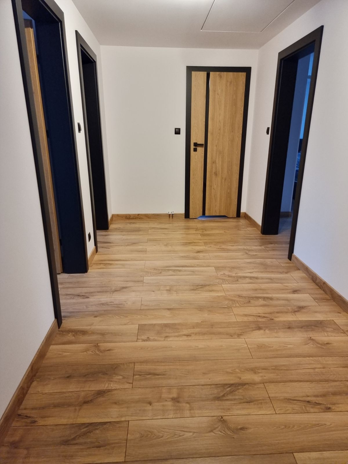 Korytarz po remoncie z drewnianą podłogą, białymi ścianami i trzema zamontowanymi drzwiami z czarnymi ościeżnicami, z czego jedne drzwi są w kolorze drewna.