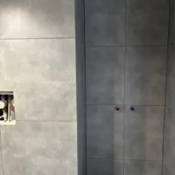 Ściana łazienki wyłożona szarymi, kwadratowymi płytkami, z widocznym otworem na rury i zawory, oraz częściowo widoczna drabina aluminiowa.