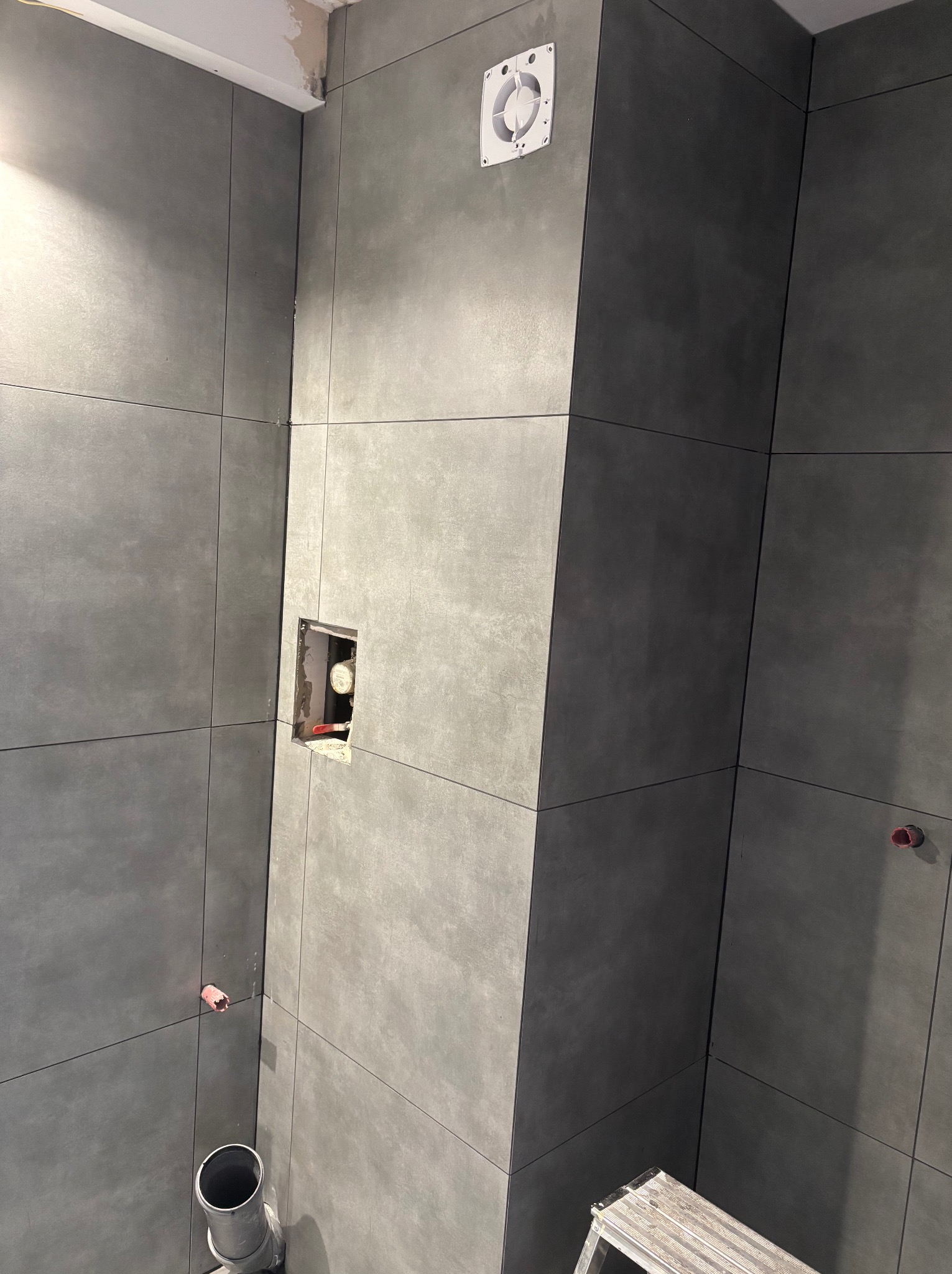 Szare, matowe płytki wielkoformatowe ułożone na ścianach łazienki, widoczny otwór pod baterię i wentylator, częściowo widoczna drabina.