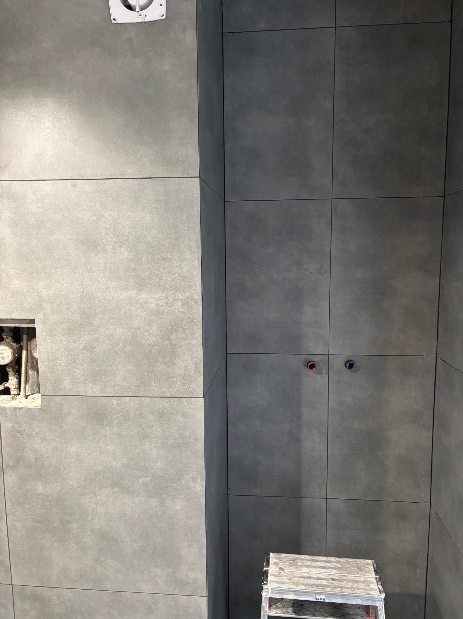 Ściana łazienki wyłożona szarymi, kwadratowymi płytkami, z widocznym otworem na rury i zawory, oraz częściowo widoczna drabina aluminiowa.