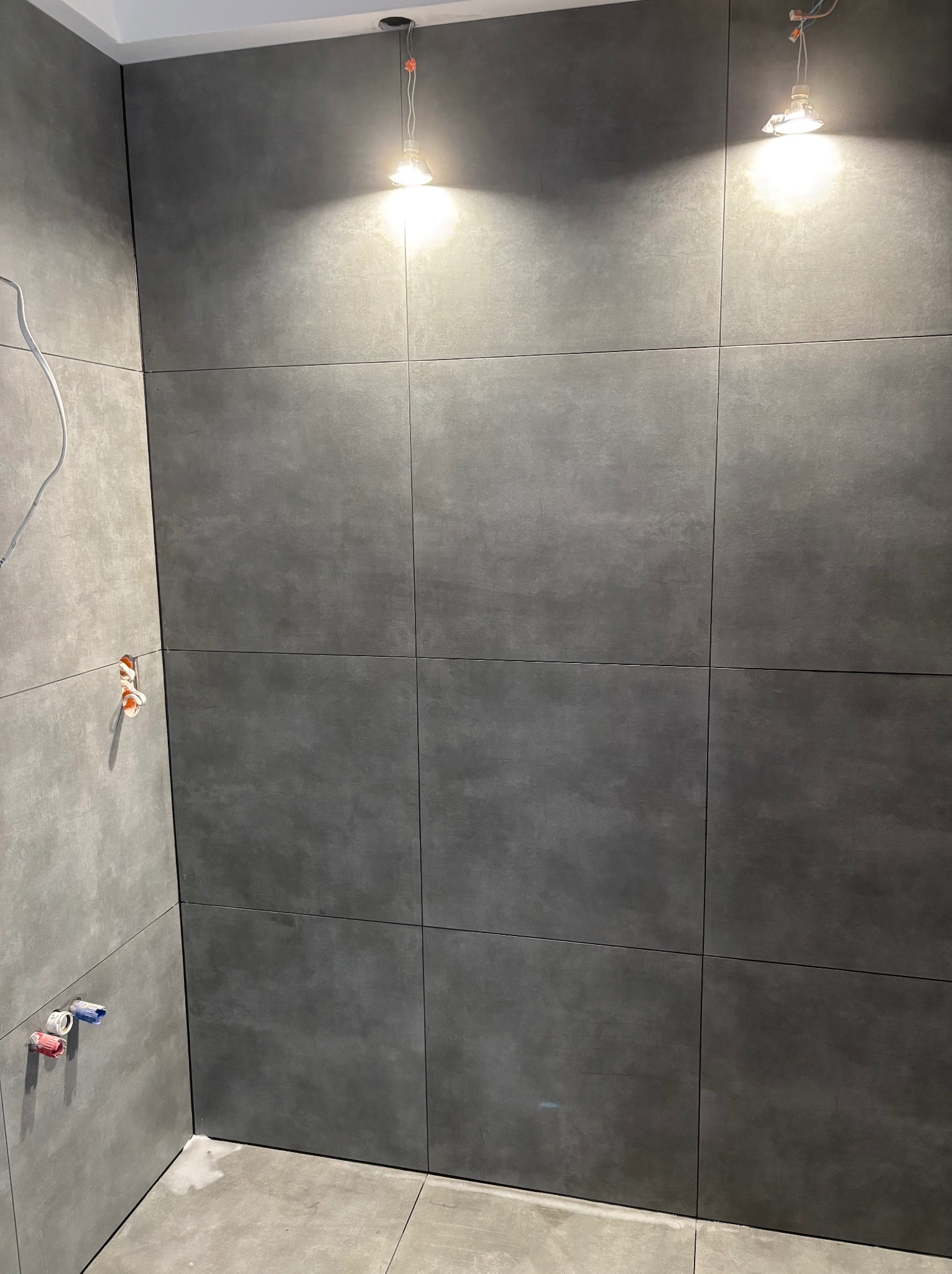 Ściana łazienki wyłożona dużymi, ciemnoszarymi płytkami, z widocznymi wystającymi przewodami i instalacją hydrauliczną, oświetlona tymczasowymi żarówkami, ukazująca etap prac wykończeniowych.