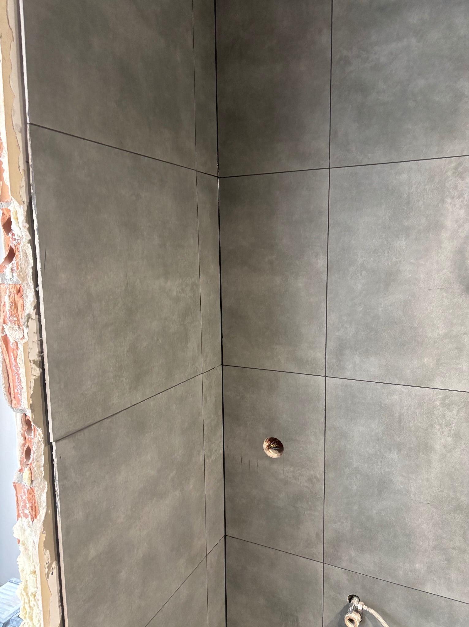 Szare płytki ceramiczne ułożone na ścianie w rogu łazienki, widoczne niedokończone wykończenie ściany z cegły oraz otwór na gniazdko elektryczne i rurę doprowadzającą wodę.