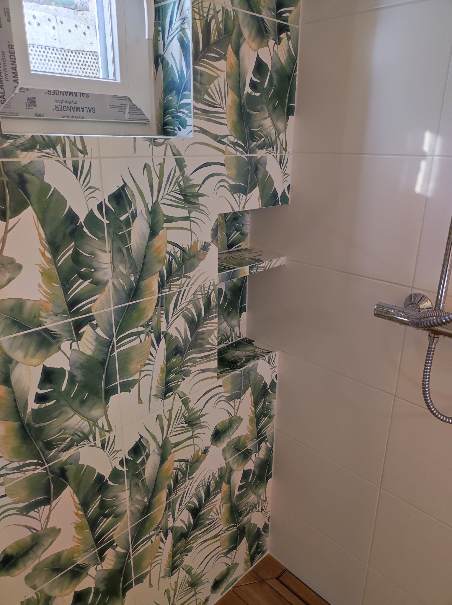 Wykończona łazienka z płytkami w tropikalne liście, wbudowanymi półkami z tego samego wzoru i widocznym fragmentem kabiny prysznicowej oraz oknem z profilem Salamander.