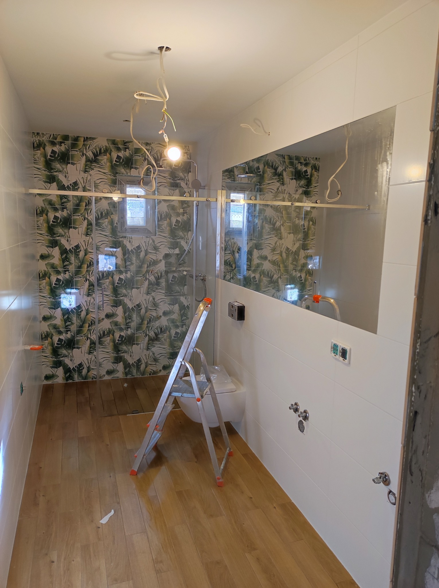 Łazienka w trakcie remontu: ułożona podłoga z paneli imitujących drewno, białe płytki na ścianach z miejscami na armaturę, kabina prysznicowa ze szklanymi drzwiami na tle ściany w tropikalne wzory,...