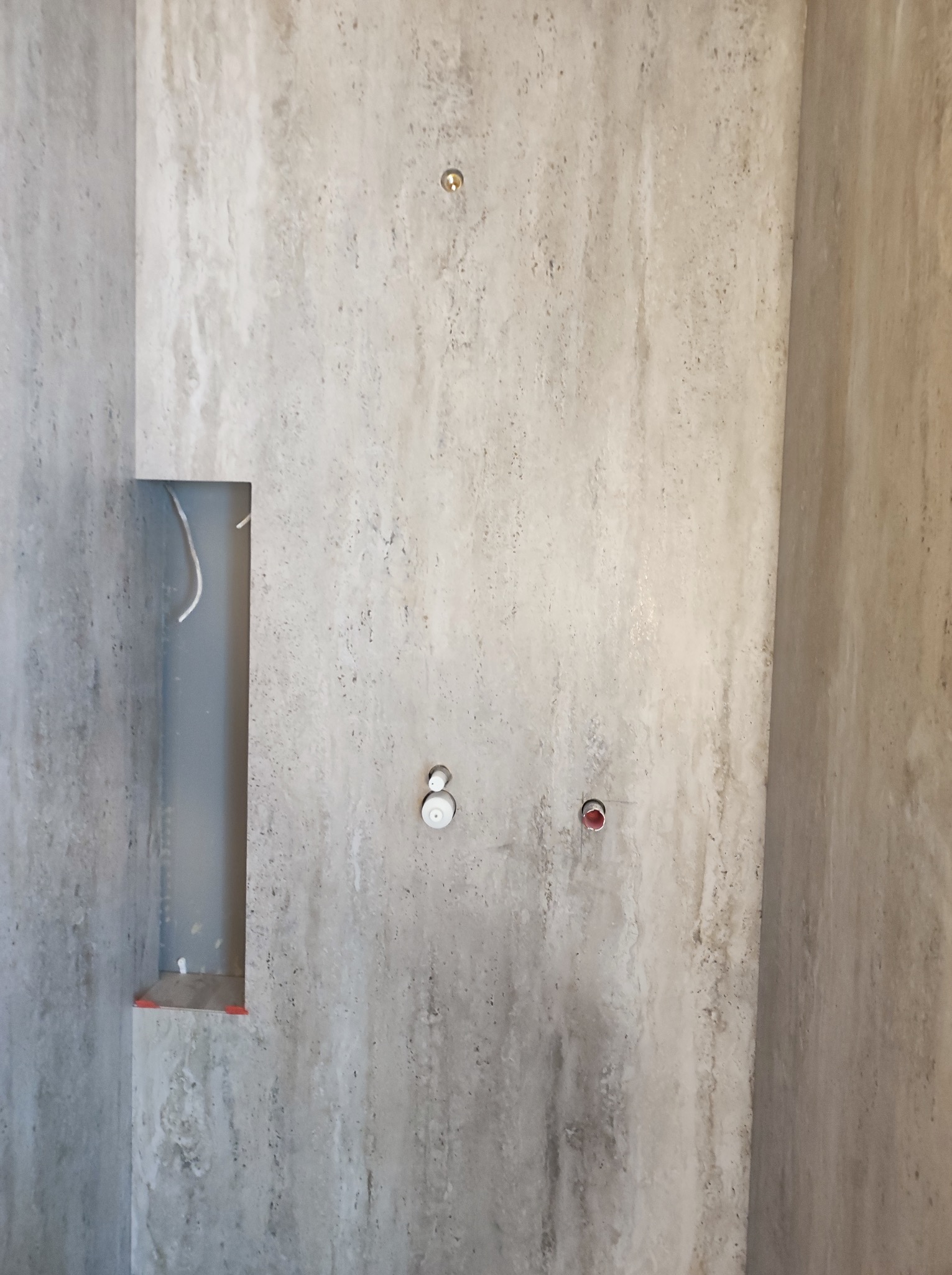 Ściana łazienki wyłożona płytkami imitującymi kamień, z widocznymi otworami na armaturę, wnęką z wystającymi przewodami i osadzonymi narożnikami zabezpieczającymi.