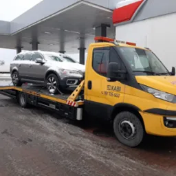 Ż&oacute;łta laweta firmy KABI z numerem telefonu 791 106 600 przewozi szary samoch&oacute;d osobowy Volvo XC70 pokryty śniegiem, na tle stacji benzynowej w zimowy dzień.