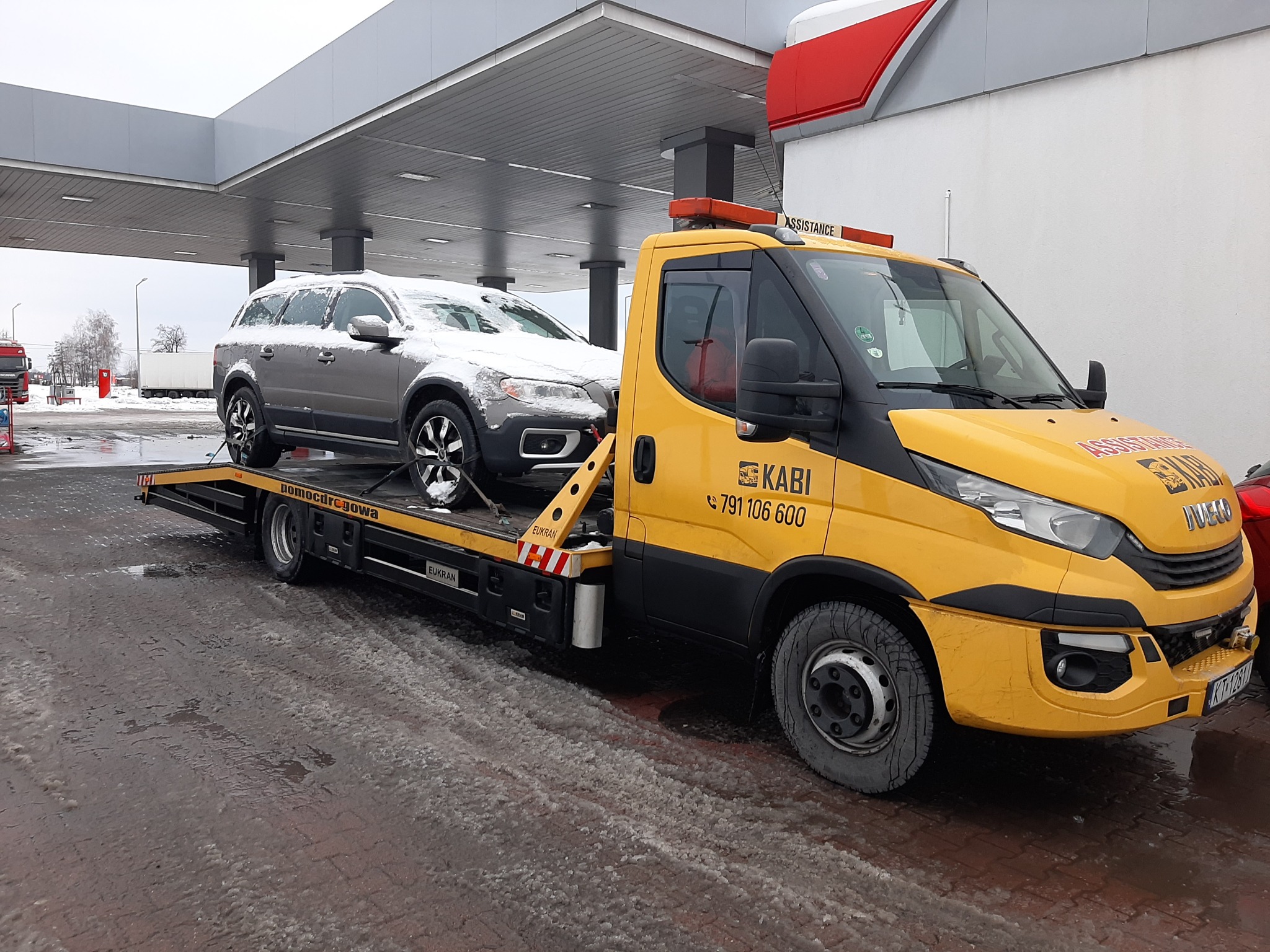 Żółta laweta firmy KABI z numerem telefonu 791 106 600 przewozi szary samochód osobowy Volvo XC70 pokryty śniegiem, na tle stacji benzynowej w zimowy dzień.