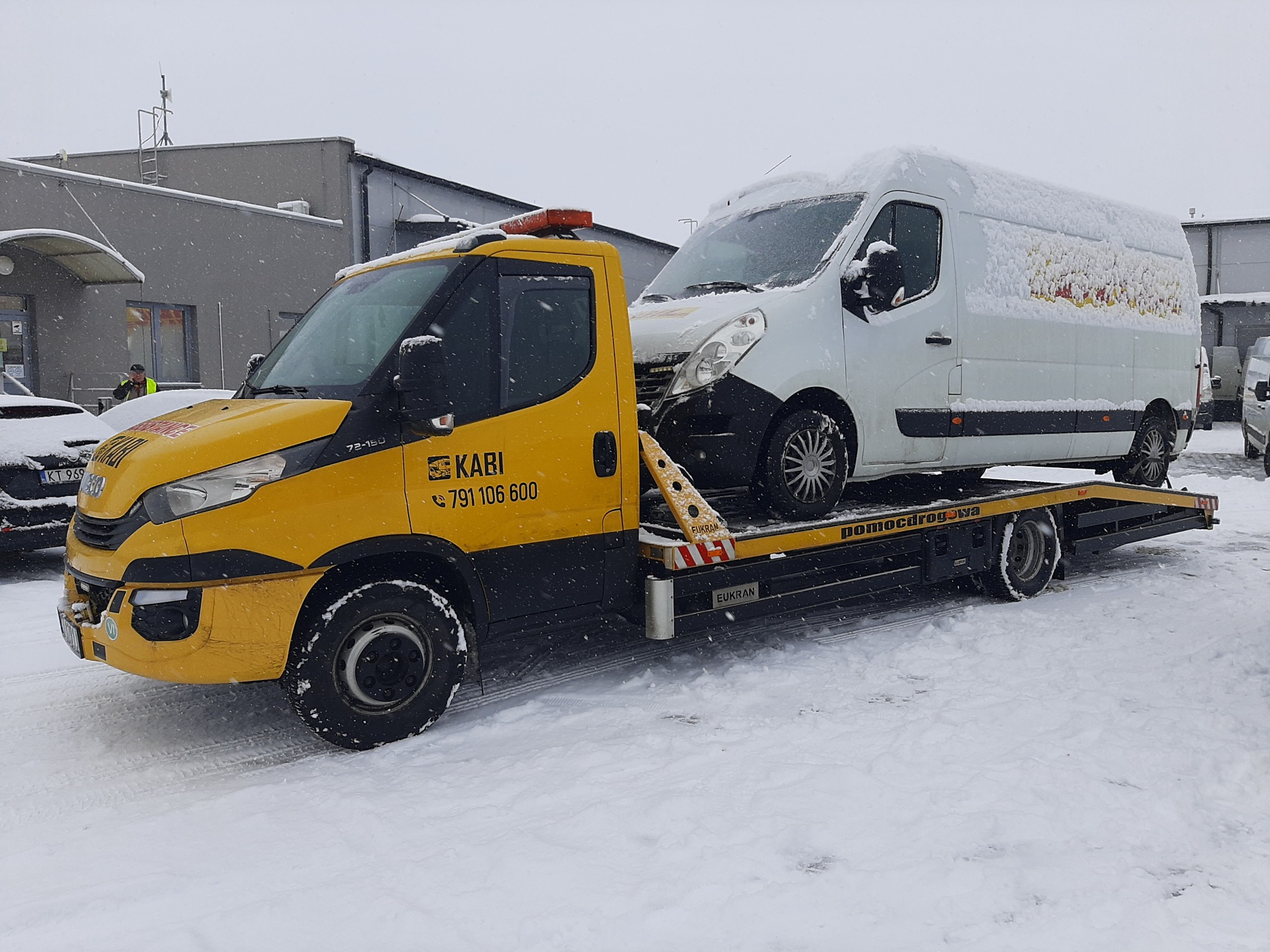 Żółto-czarna laweta KABI z białym vanem pokrytym śniegiem na platformie, zimowy krajobraz.