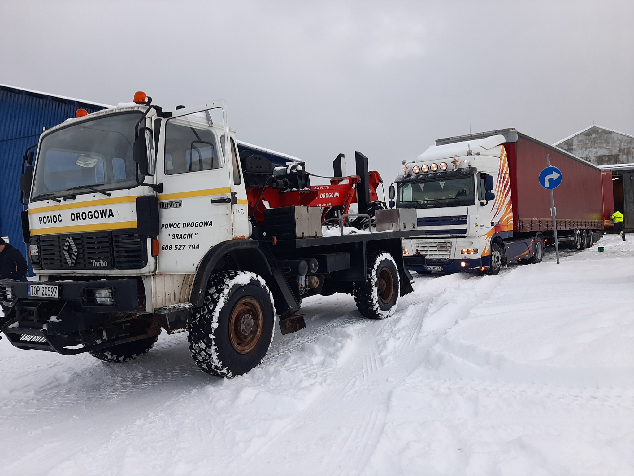 Ciężarówka pomocy drogowej Renault z dźwigiem holuje inną ciężarówkę w zimowych warunkach, widoczny śnieg i budynki w tle.
