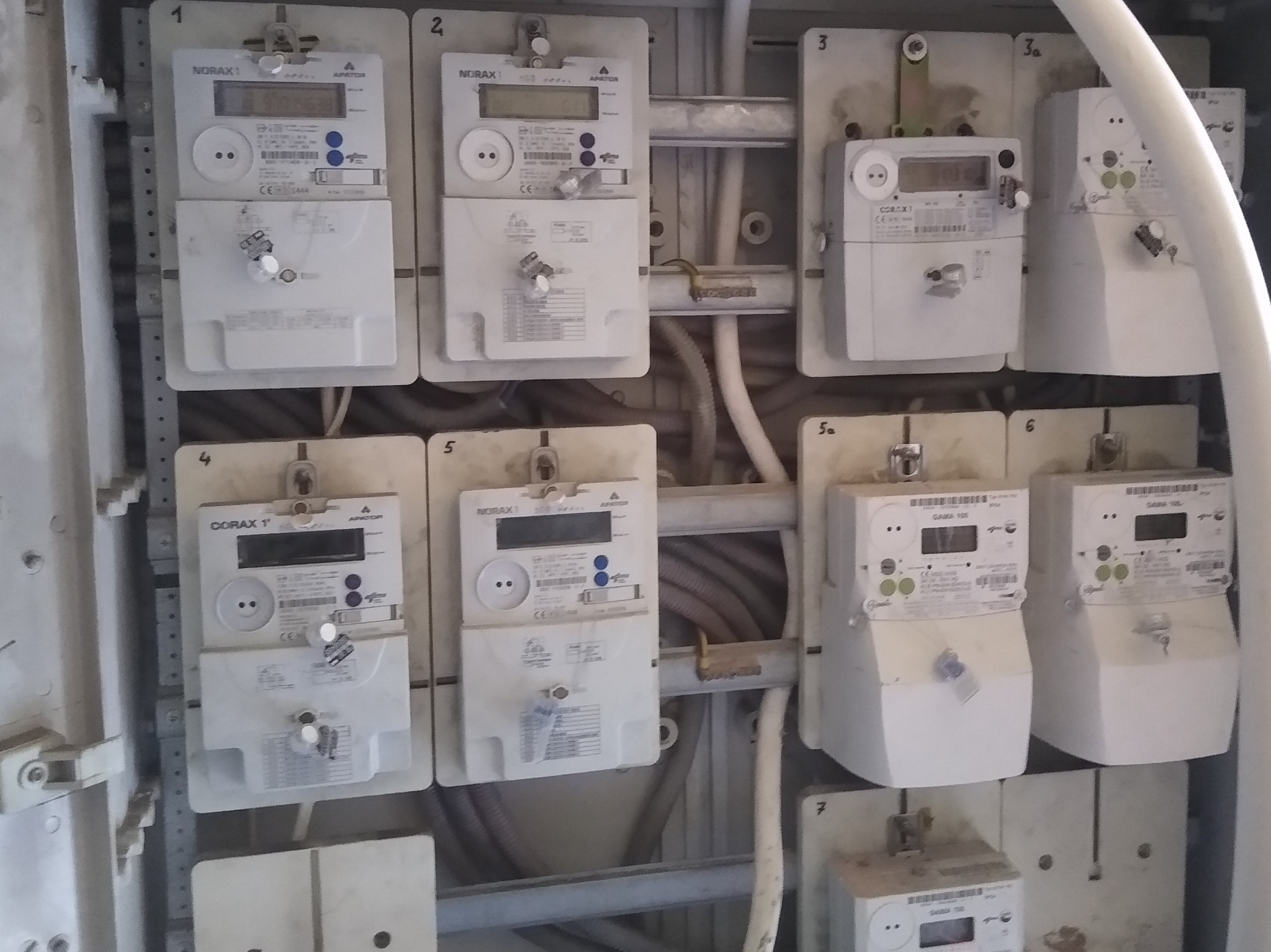 Szafka elektryczna z zamontowanymi licznikami energii elektrycznej różnych typów (NORAX, CORAX, GAMA) z widocznymi przewodami i numerami identyfikacyjnymi.