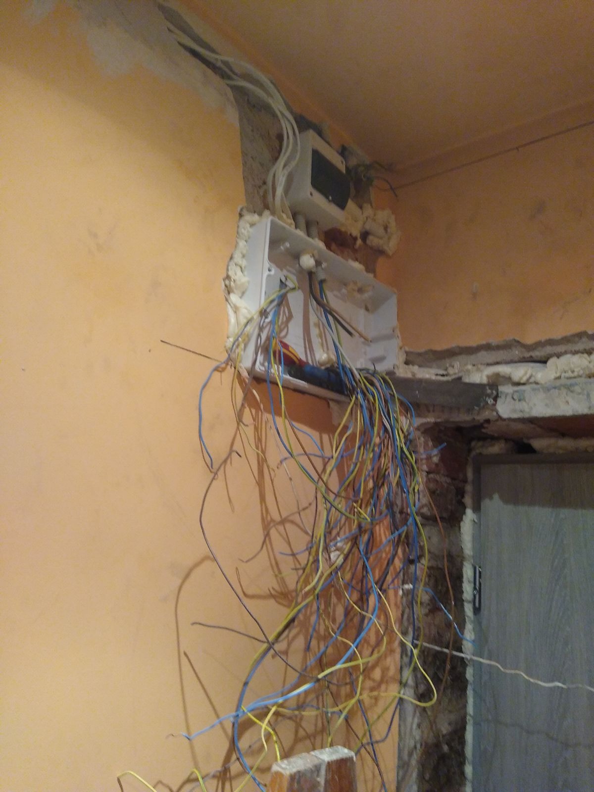 Otwarta puszka elektryczna na ścianie w trakcie prac, z wystającymi przewodami w kolorach niebieskim, żółtym i brązowym, zabezpieczona pianką montażową, widoczne fragmenty surowej ściany i cegły.