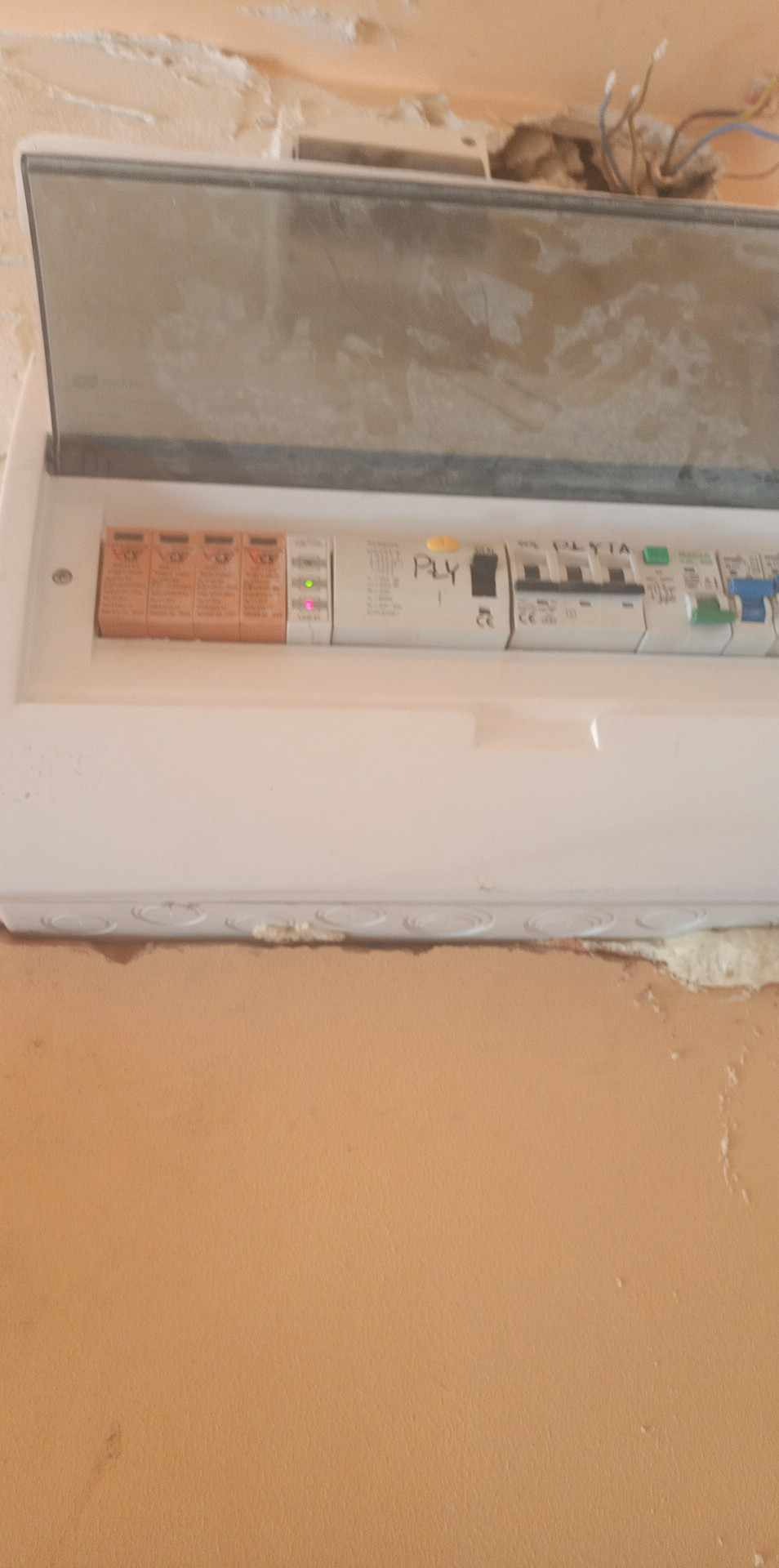 Otwarta, biała skrzynka elektryczna z bezpiecznikami i zabezpieczeniami różnicowoprądowymi, zamontowana na pomarańczowej ścianie z widocznymi śladami po instalacji, z wystającymi przewodami u góry.