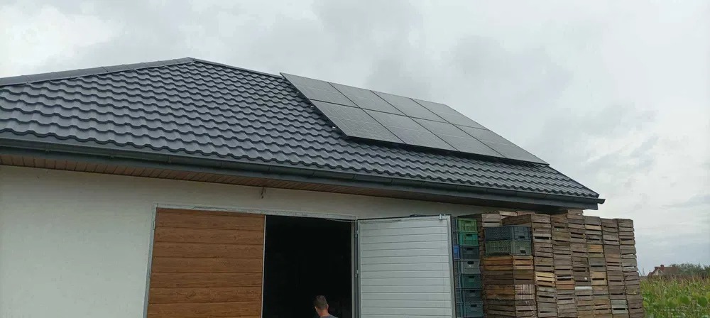 Panele słoneczne na dachu budynku gospodarczego z szarą blachodachówką, obok otwarte drzwi i sterta drewnianych skrzynek.