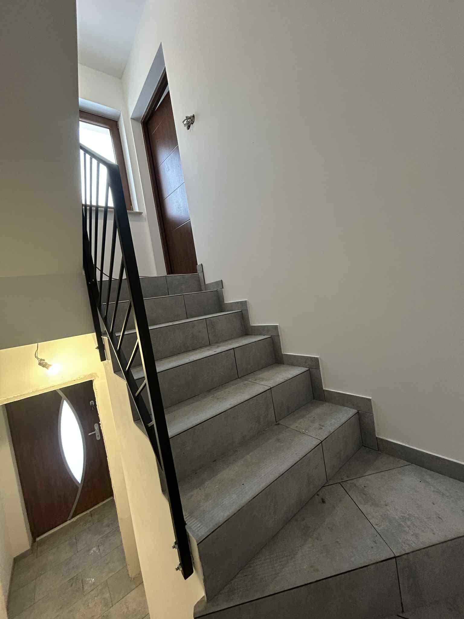 Wykończone schody wewnątrz budynku z szarymi płytkami i czarną balustradą. Widoczne drzwi wejściowe i wewnętrzne. Minimalistyczny styl wykończenia.