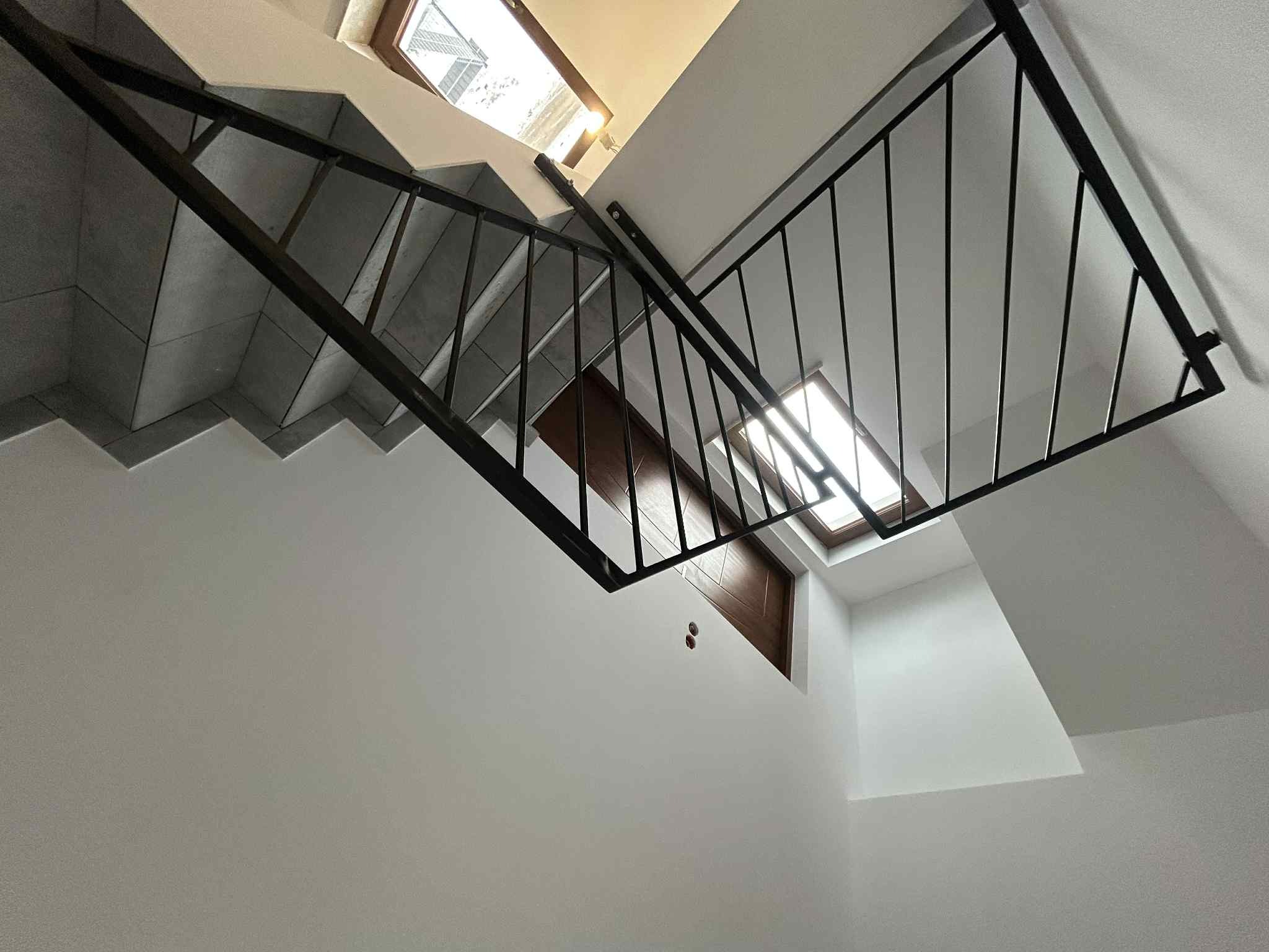 Nowoczesne schody z czarną, metalową balustradą i szarymi stopniami, widziane z dołu, z oknem dachowym w tle. Minimalistyczny design.