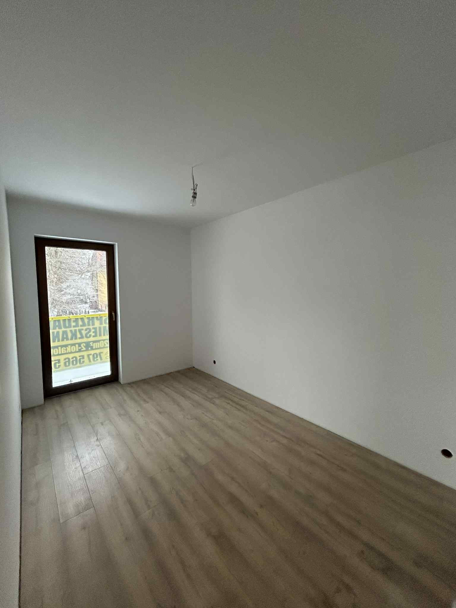 Jasne panele podłogowe w pustym pokoju z białymi ścianami i oknem z brązową ramą. Widoczny surowy kabel oświetleniowy. Minimalistyczne wykończenie wnętrza.