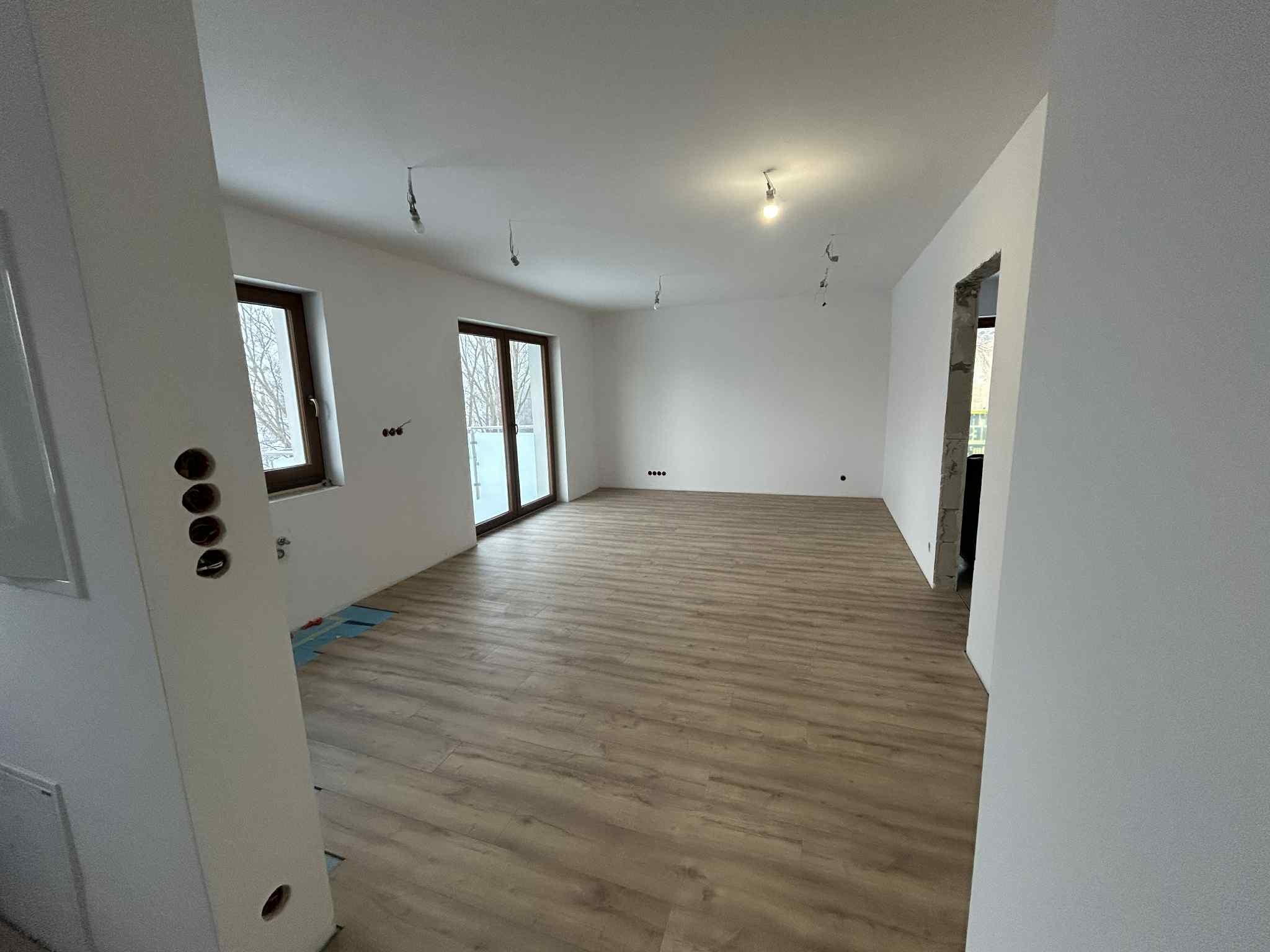 Jasne wnętrze po remoncie: panele podłogowe imitujące drewno, świeżo pomalowane białe ściany, okna z widokiem na zewnątrz, przygotowane punkty oświetleniowe na suficie.