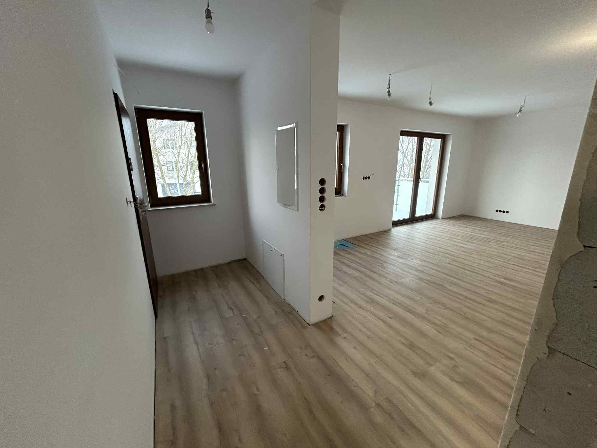 Jasne, przestronne wnętrze po remoncie z panelami podłogowymi, białymi ścianami i oknami z ciemnymi ramami. Widoczne gniazdka elektryczne i surowe wykończenie przy filarze.