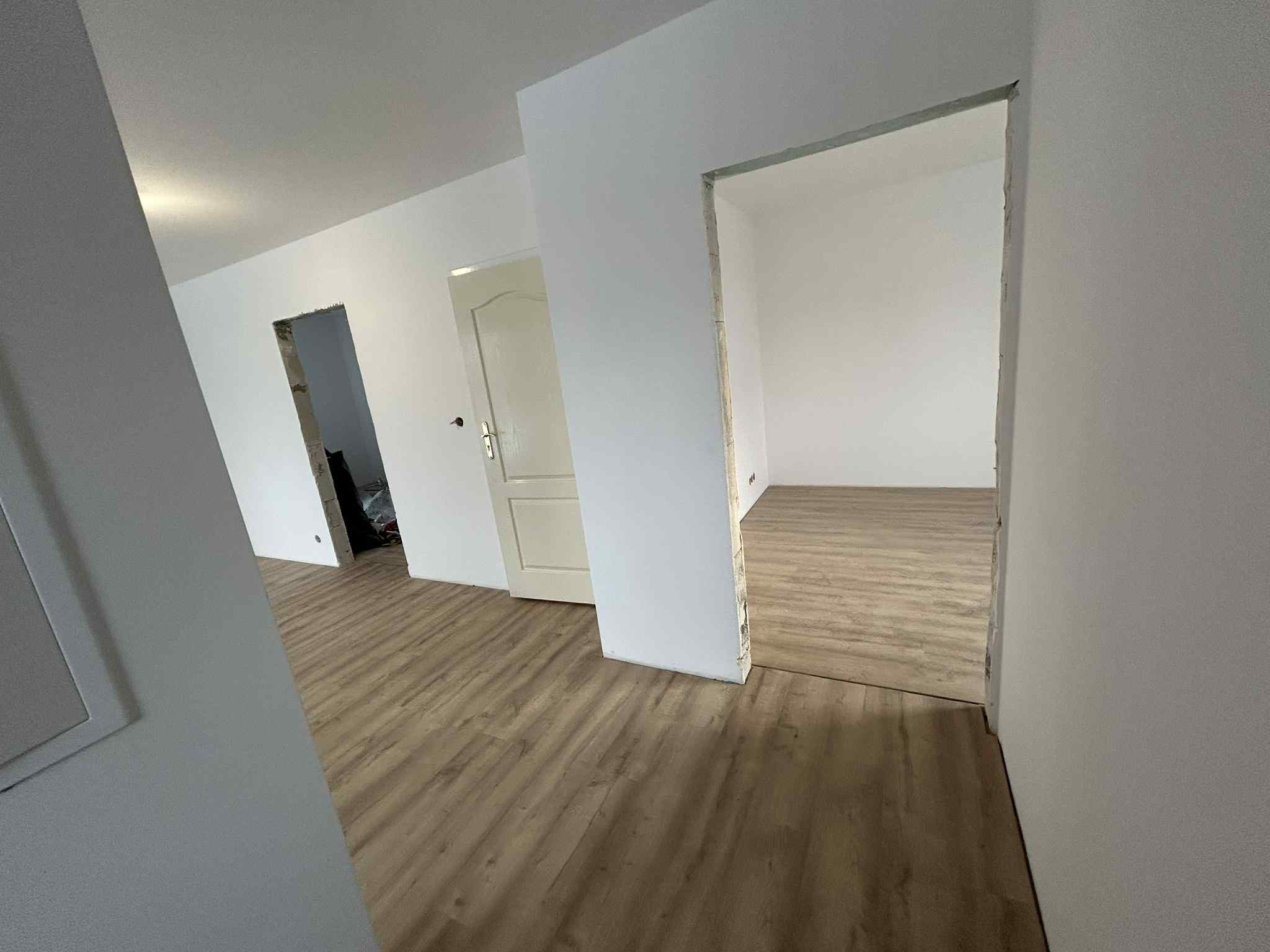 Jasne wnętrze w trakcie wykończenia: świeżo położona podłoga, białe ściany, widoczne otwory drzwiowe i fragmenty instalacji. Minimalistyczny styl, surowy stan.