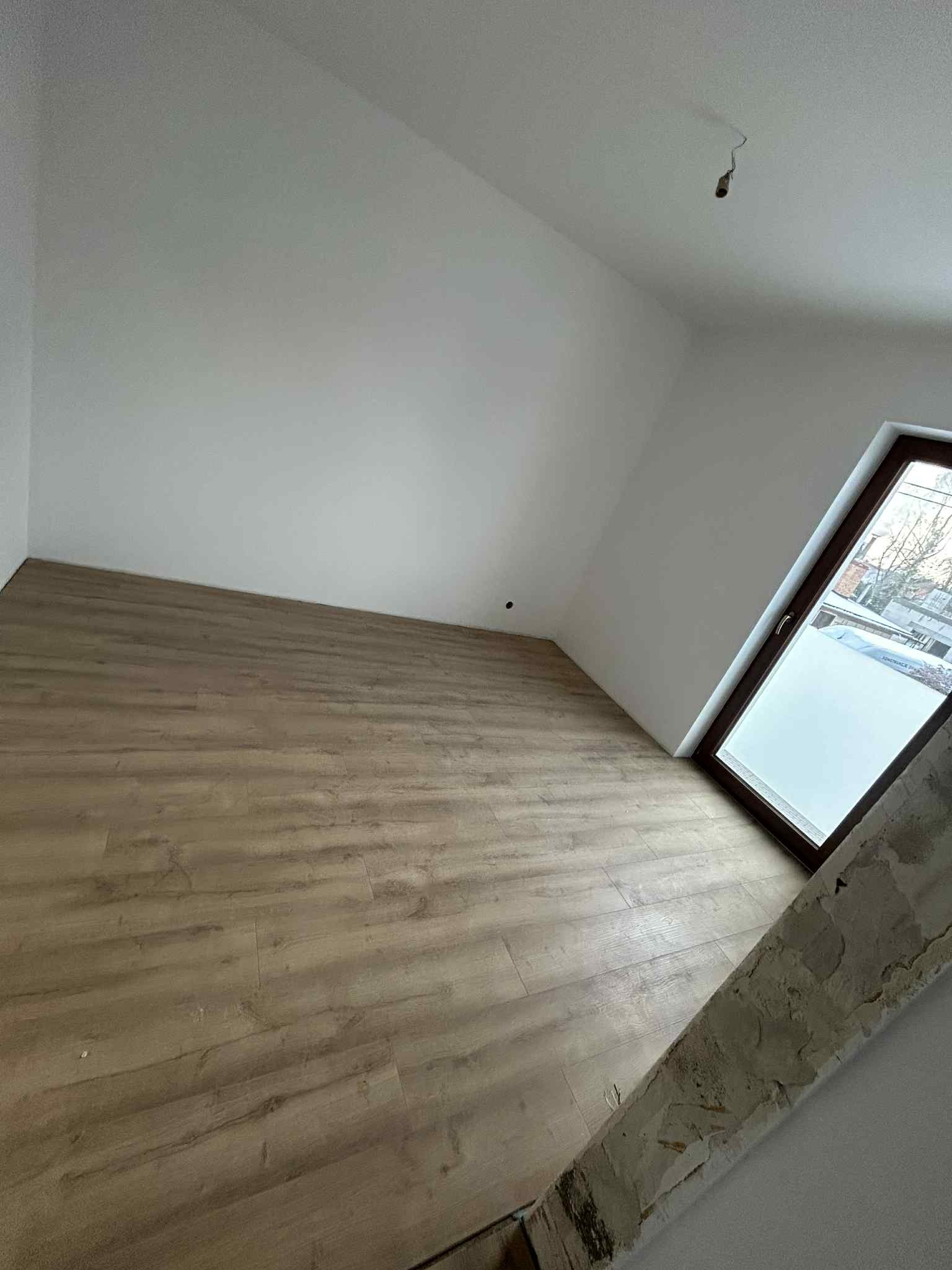 Pokój na poddaszu z drewnianą podłogą i oknem. Ściany białe, widoczne ślady po remoncie przy oknie. Ujęcie z dołu, podkreślające skos dachu.