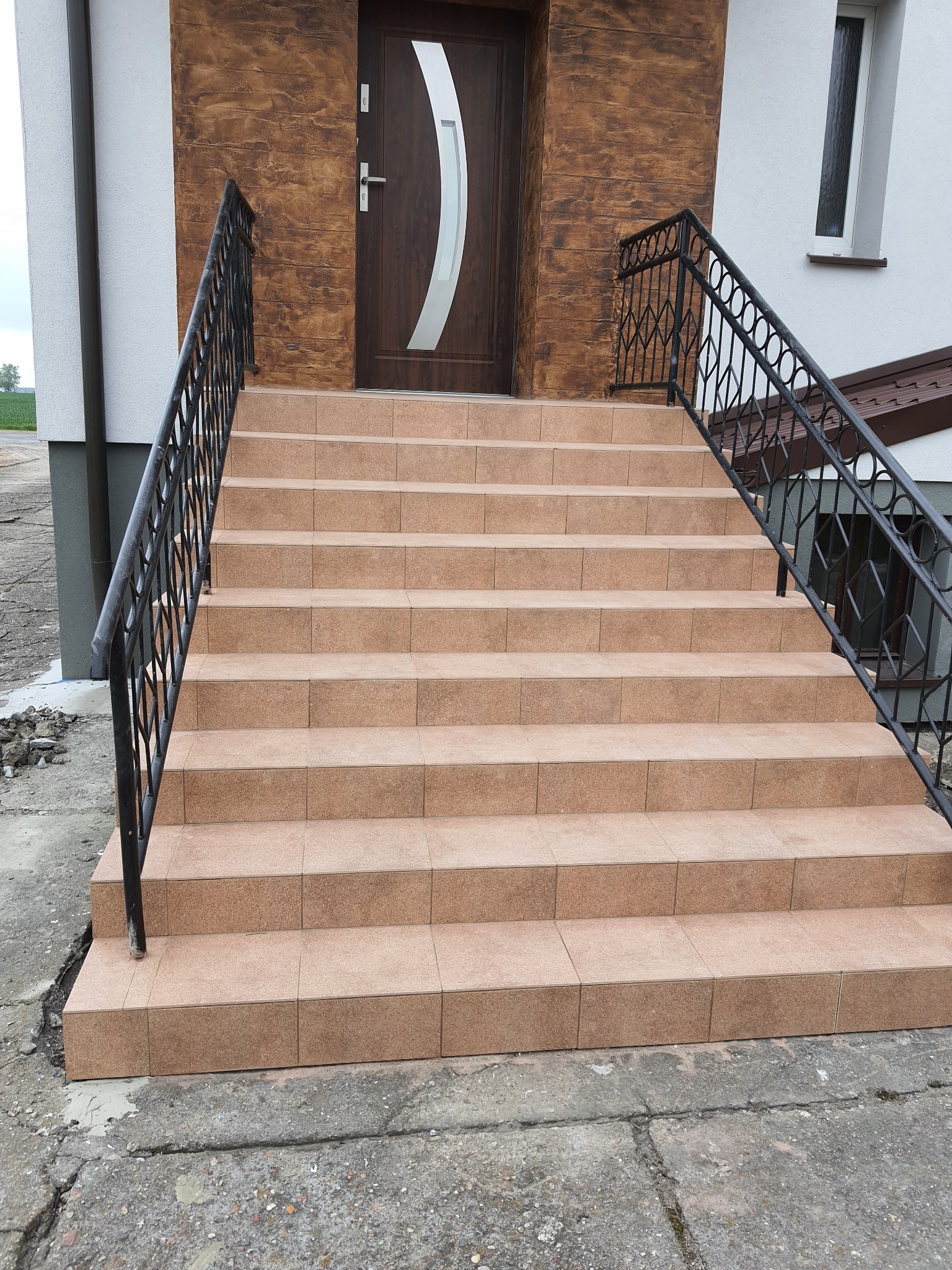 Zewnętrzne schody wejściowe do domu, wyłożone beżowymi płytkami, z czarną, metalową balustradą o ozdobnym wzorze, prowadzące do brązowych drzwi z białym akcentem, na tle białej ściany i fragmentu...